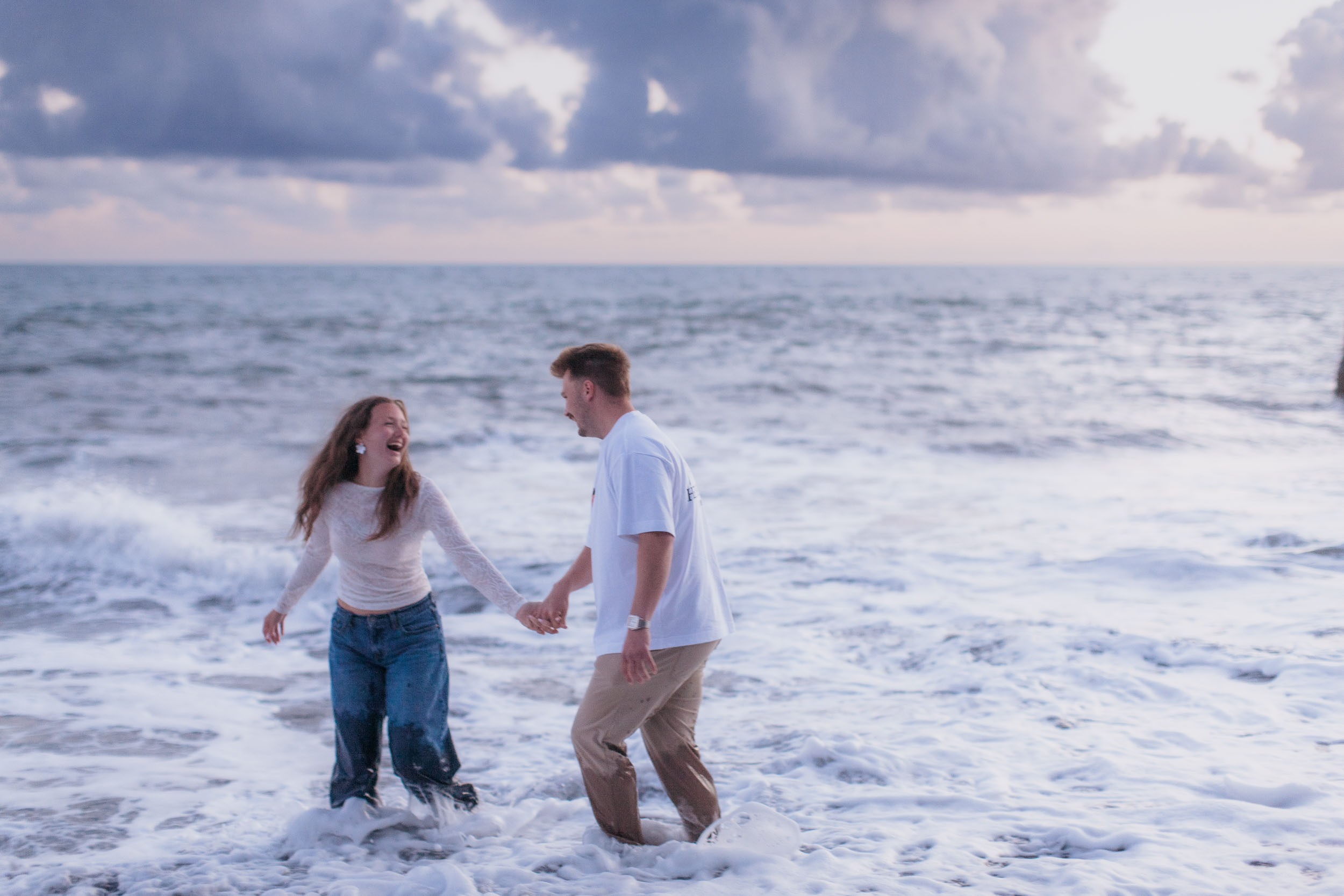 Standing in the surf holding hands, she laughs with her head thrown back, heavy cloud above the horizon, Praia Formosa, Madeira