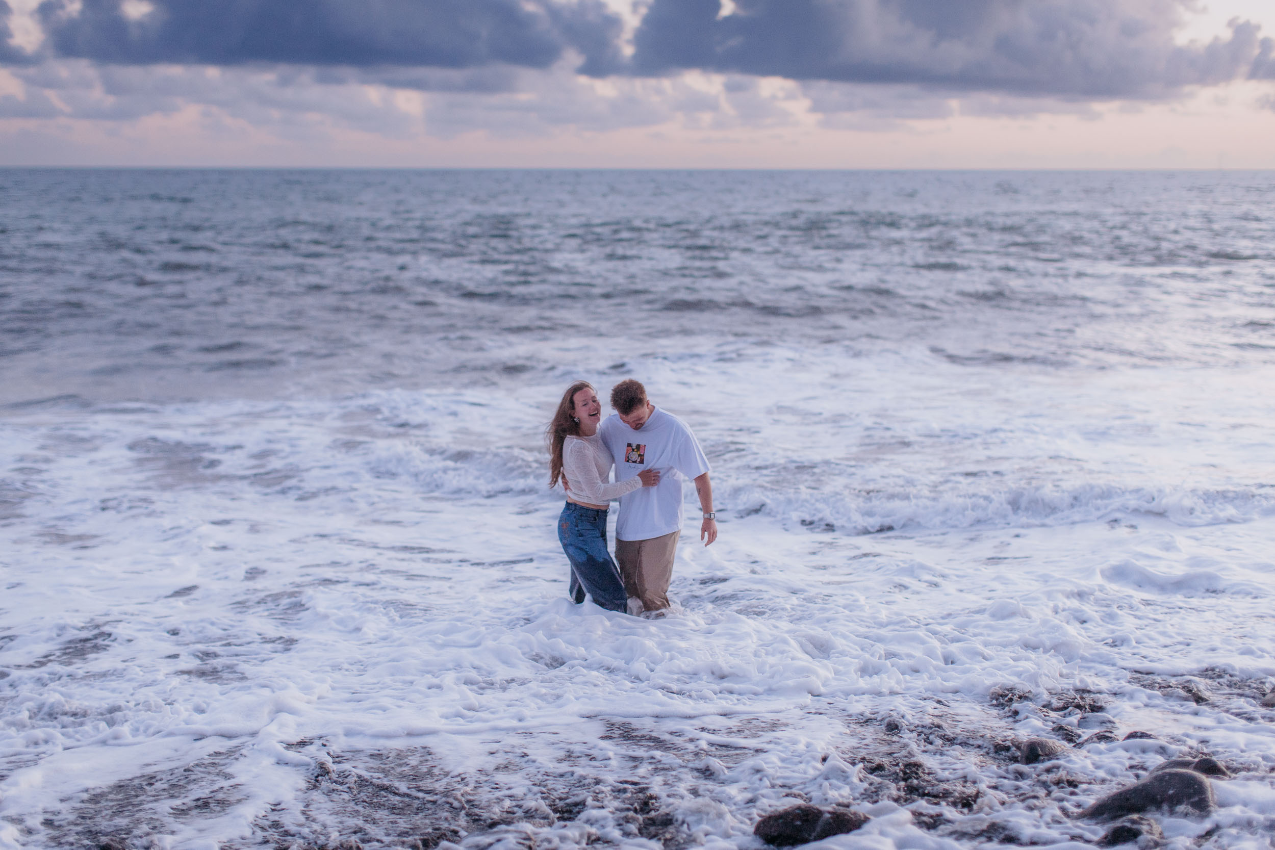 Wide frame. The two of them small in the white foam, laughing, the whole beach empty around them, Praia Formosa, Madeira