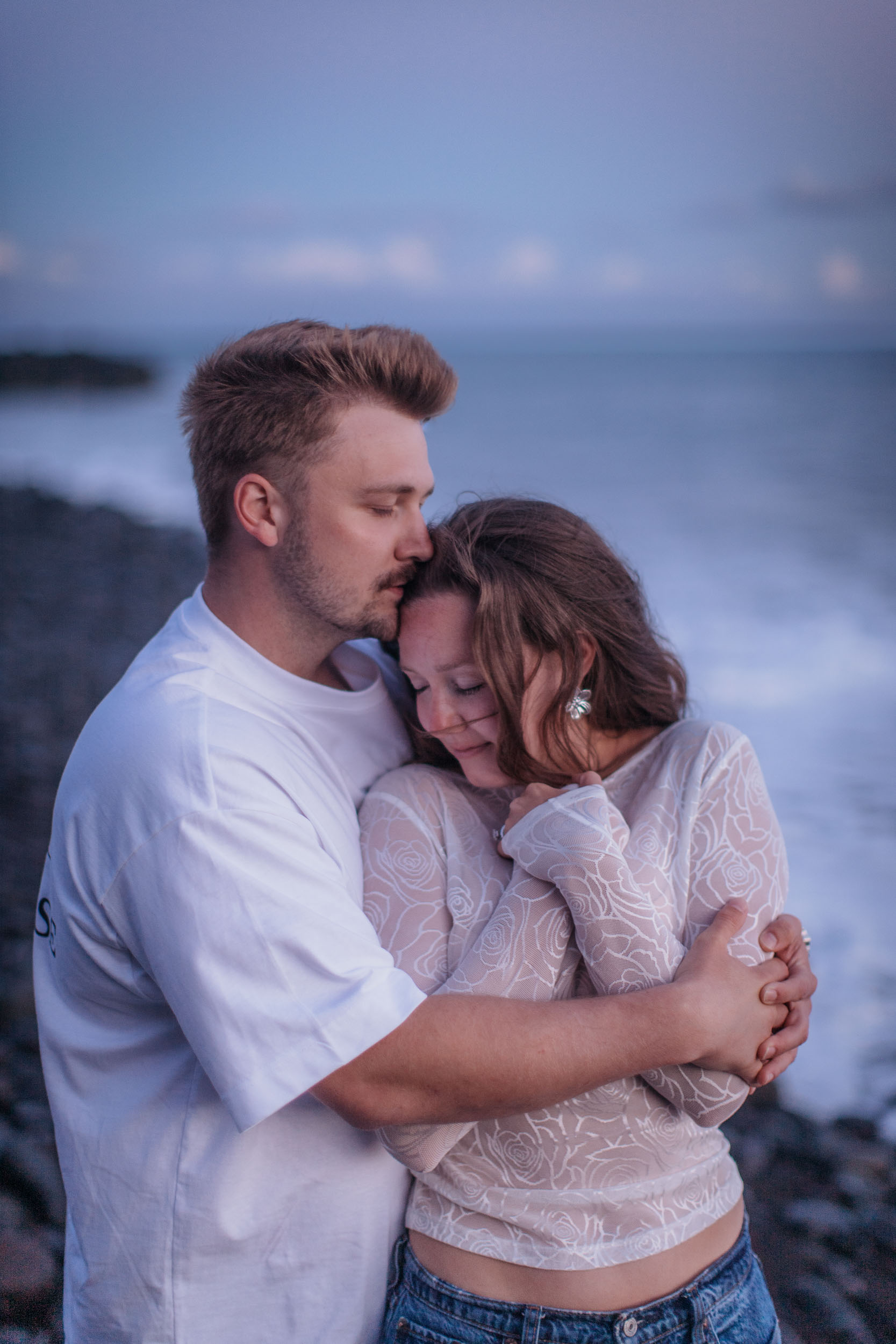 Vertical frame. He holds her from behind and kisses the top of her head, both with eyes closed at blue hour, Praia Formosa, Madeira