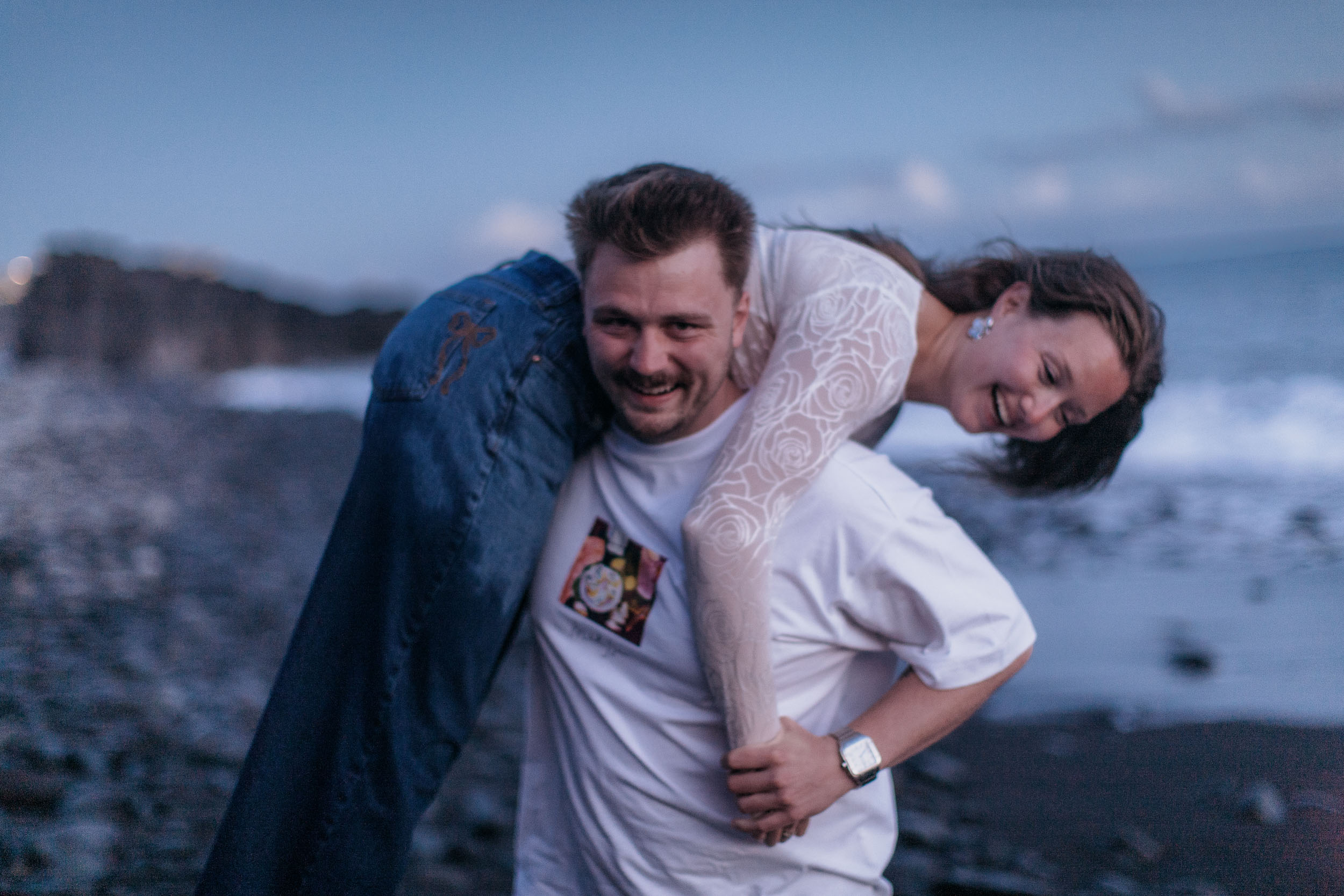He carries her over his shoulder and both are laughing, the beach going dark behind them, Praia Formosa, Madeira