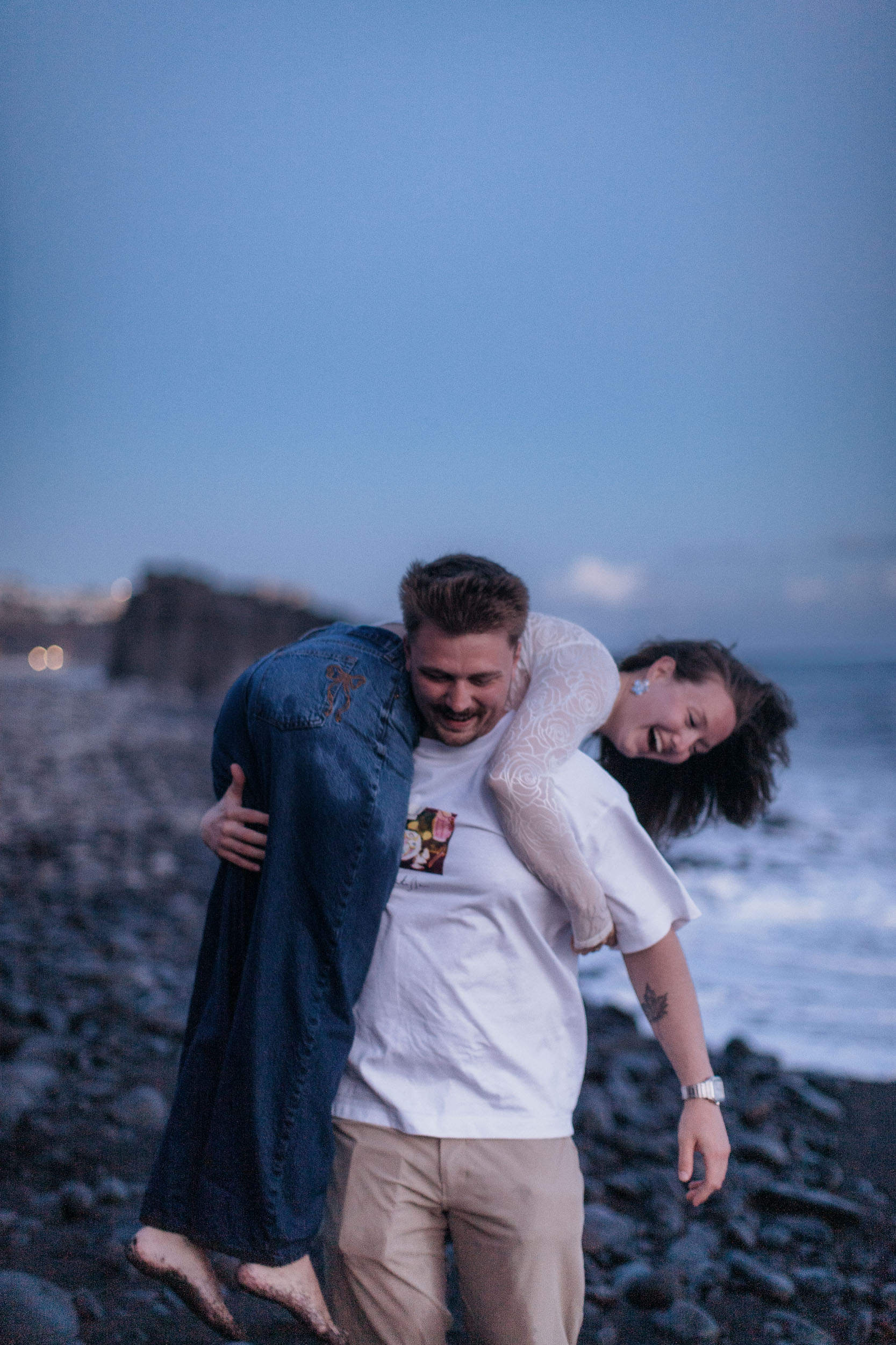 Vertical frame. Carrying her over his shoulder along the stones at blue hour, both laughing, lights of the village behind, Praia Formosa, Madeira