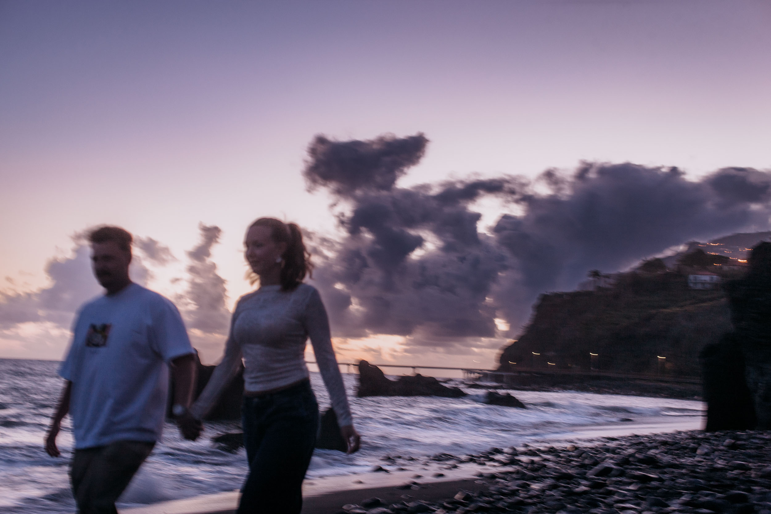Rylie and Abby walking along Praia Formosa shoreline at blue hour, soft motion blur, purple cloud sky and distant Funchal cliffs, Madeira