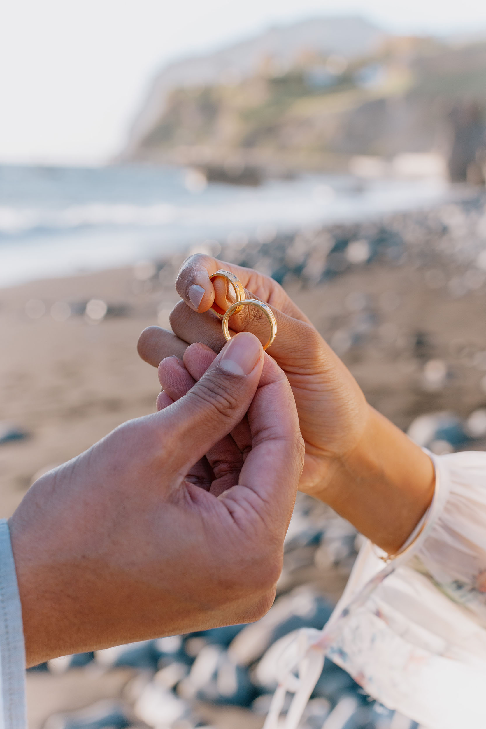 Two gold engagement rings held together at Praia Formosa beach, Funchal cliffs visible in the background, Madeira