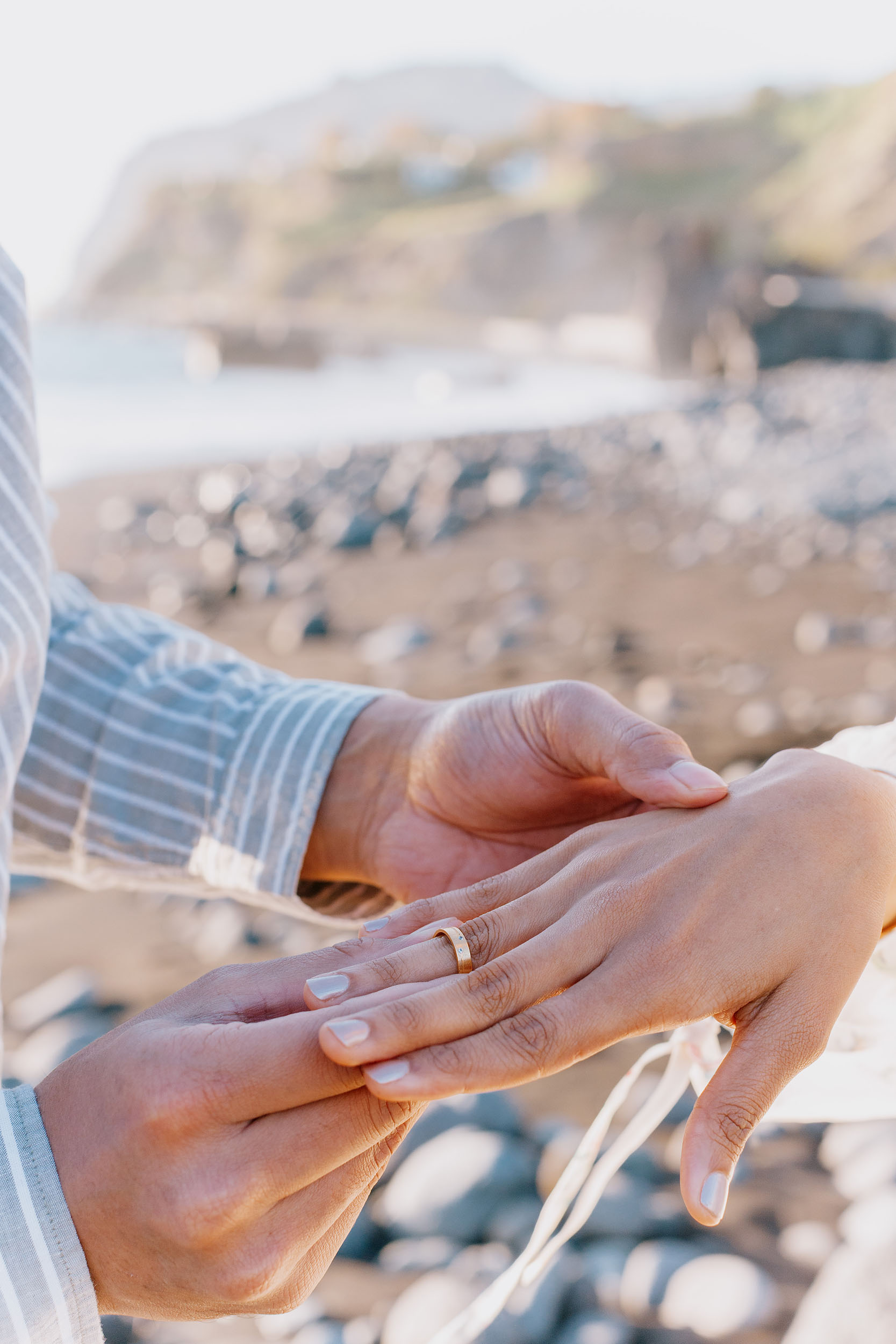 Saket sliding an engagement ring onto Sneha's finger on volcanic rocks at Praia Formosa, Funchal cliffs behind, Madeira