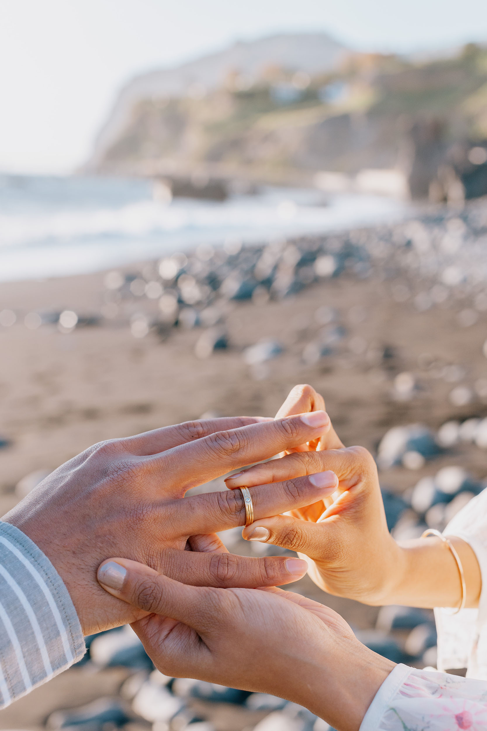 Saket placing ring on Sneha's finger, golden sunset light, Funchal cliffs soft in the bokeh background, Praia Formosa, Madeira