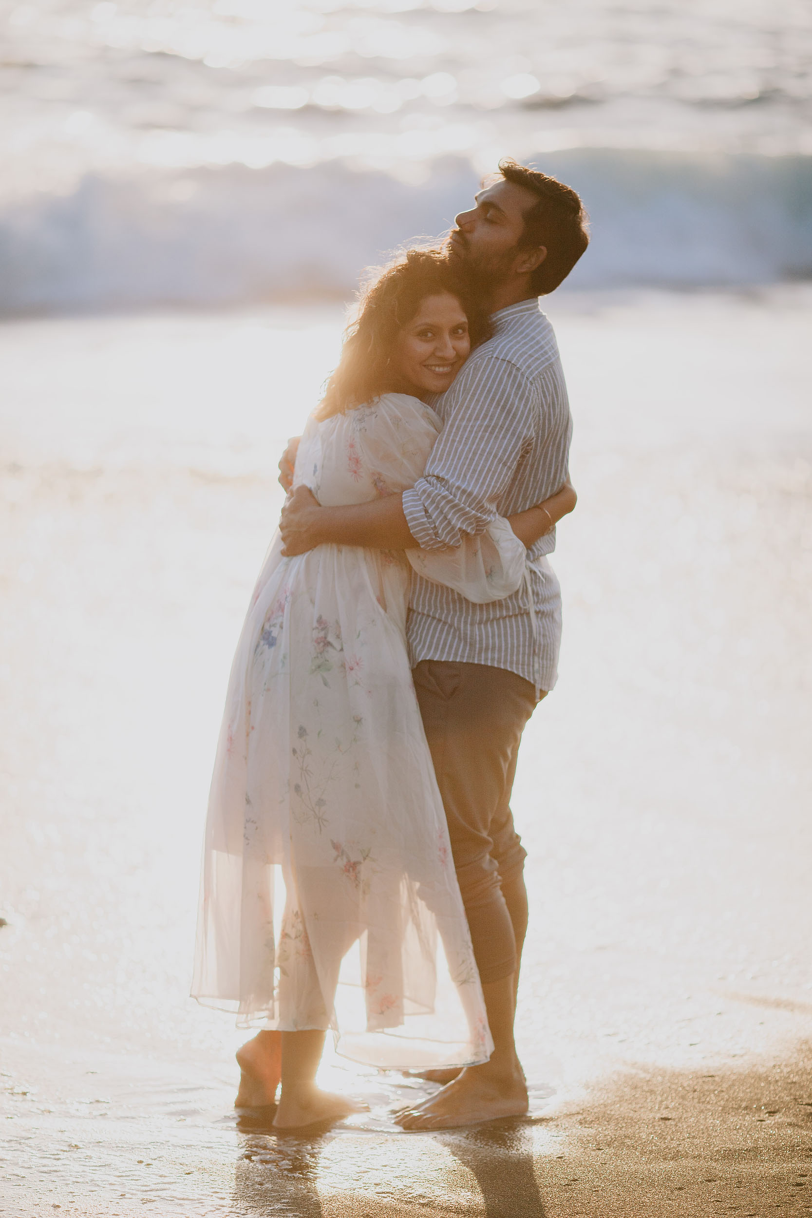 Sneha embracing Saket from behind, both figures backlit by strong sunset flare, Praia Formosa shoreline, Madeira