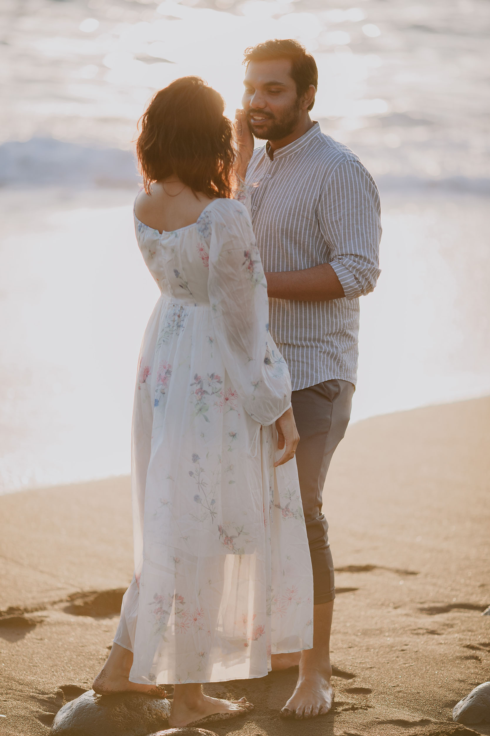 Saket gently touching Sneha's face at Praia Formosa beach, warm backlit evening light, Funchal, Madeira