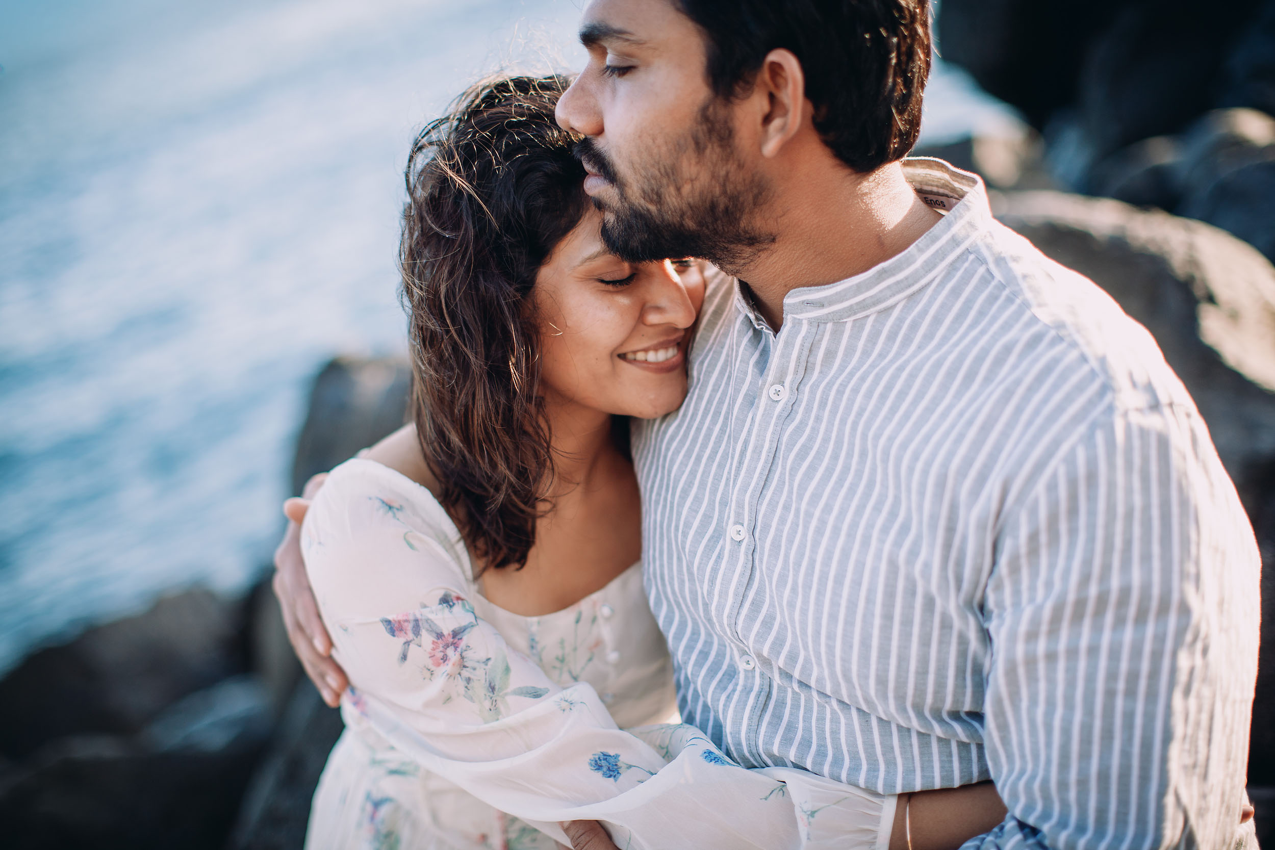 Saket kissing Sneha's temple while she smiles, both sitting on volcanic rocks at Praia Formosa, Atlantic ocean behind, Madeira