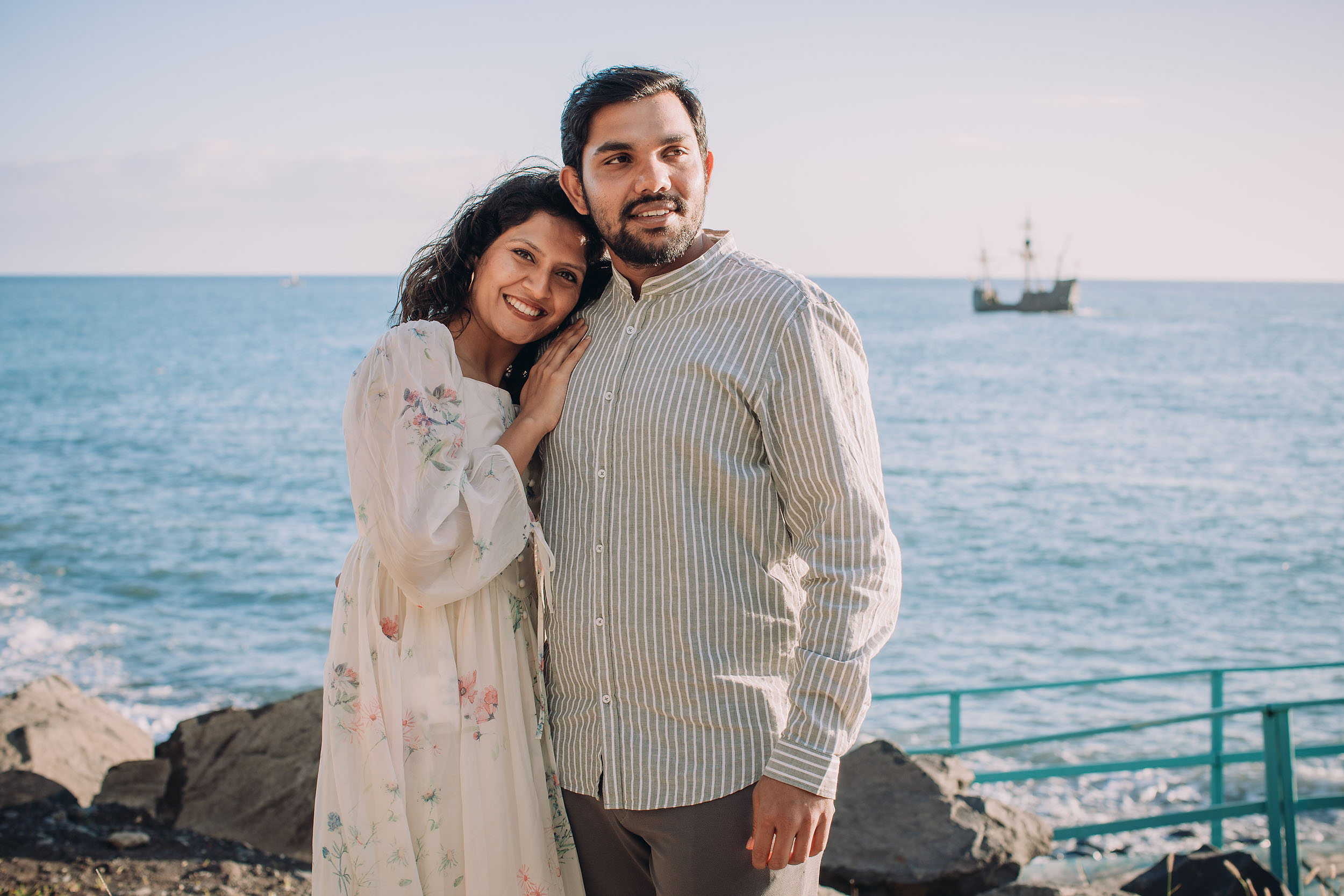 Sneha leaning smiling against Saket, historic tall ship visible in Funchal harbour behind them, Praia Formosa, Madeira