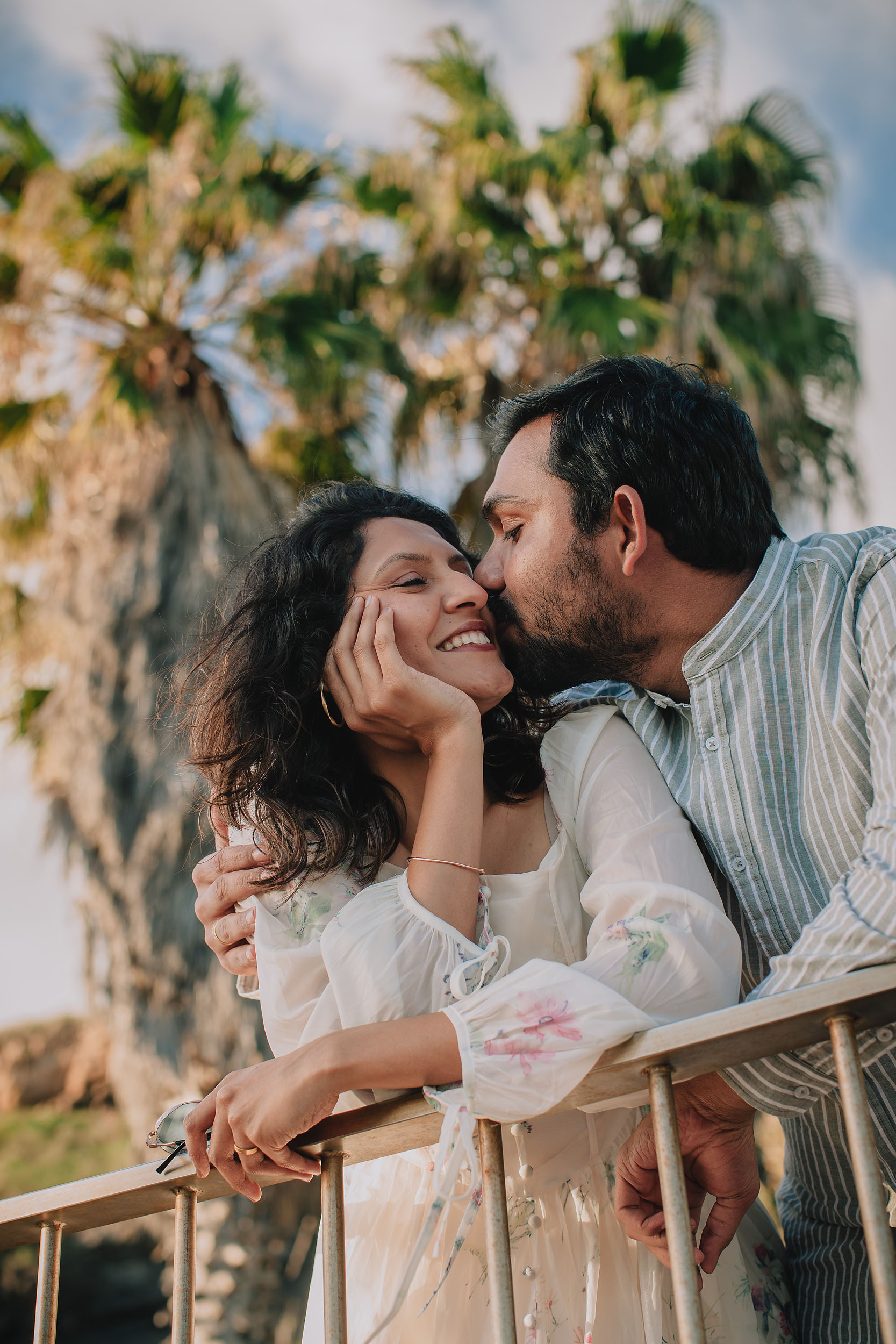 Saket kissing Sneha on the cheek as she smiles, large palm trees and Funchal promenade railing behind them, Madeira