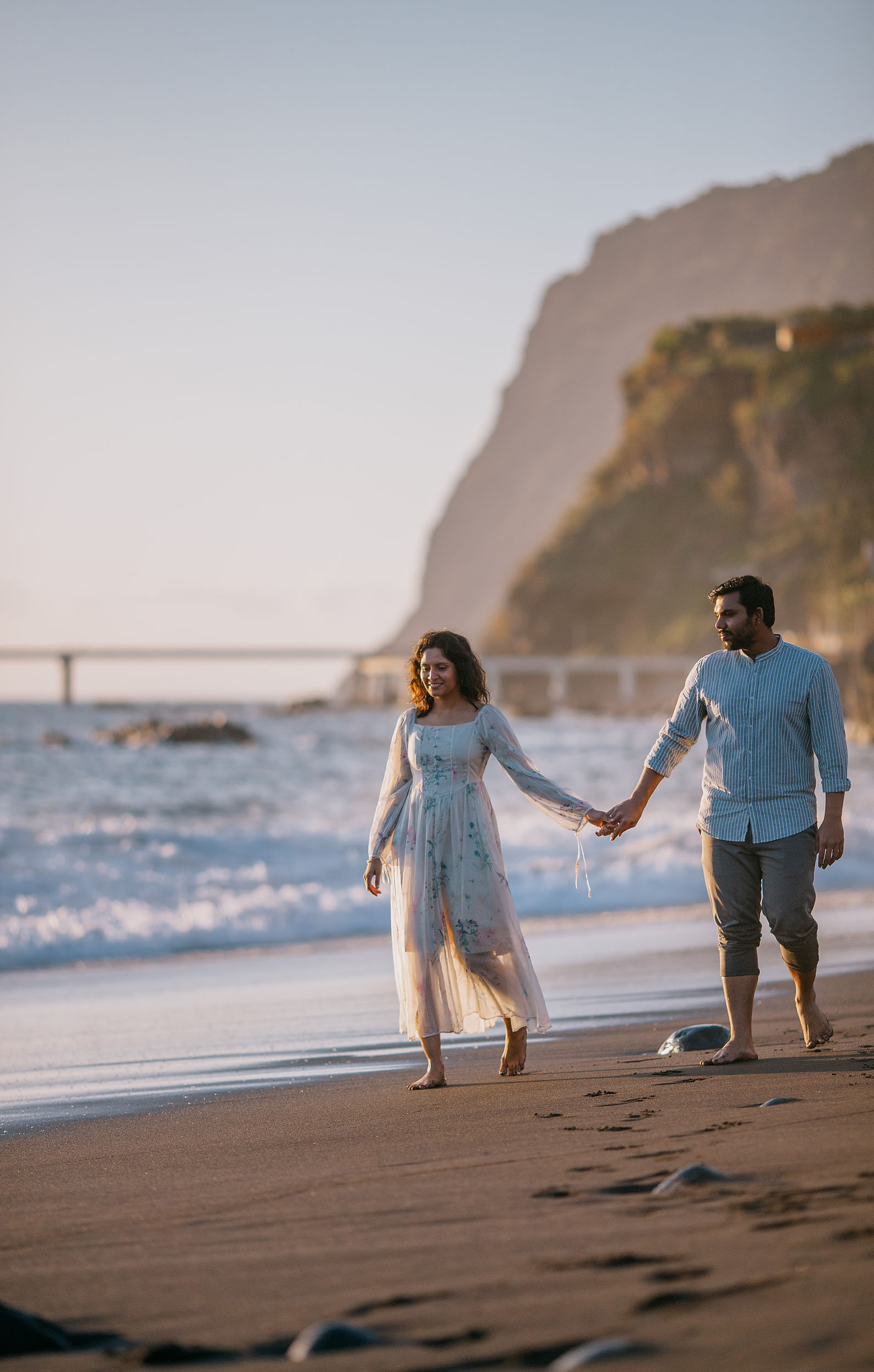 Saket and Sneha walking hand in hand along Praia Formosa beach, Funchal cliffs in the background, warm evening light, Madeira
