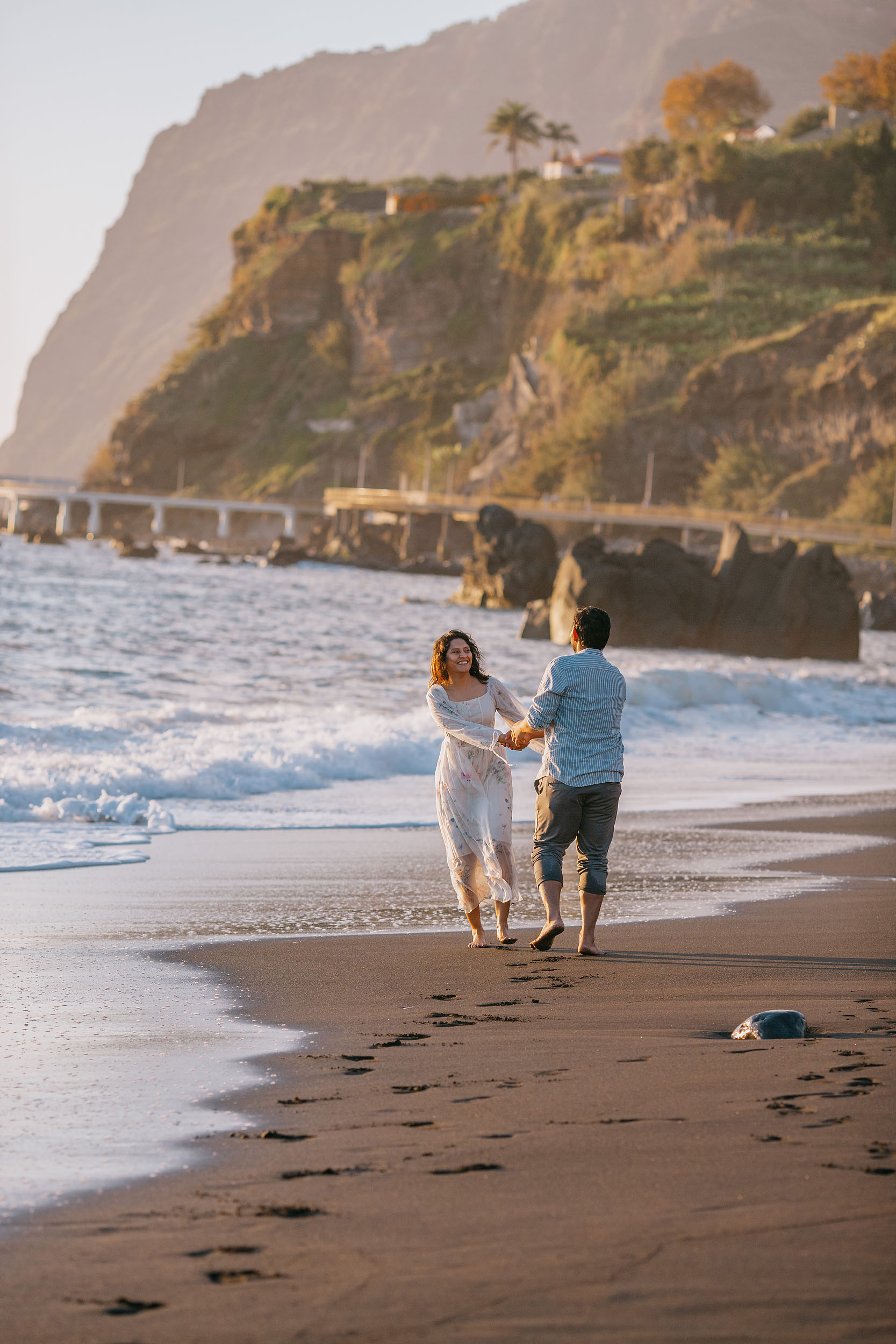 Saket and Sneha running hand in hand toward the camera along Praia Formosa shoreline, Funchal cliffs behind, Madeira