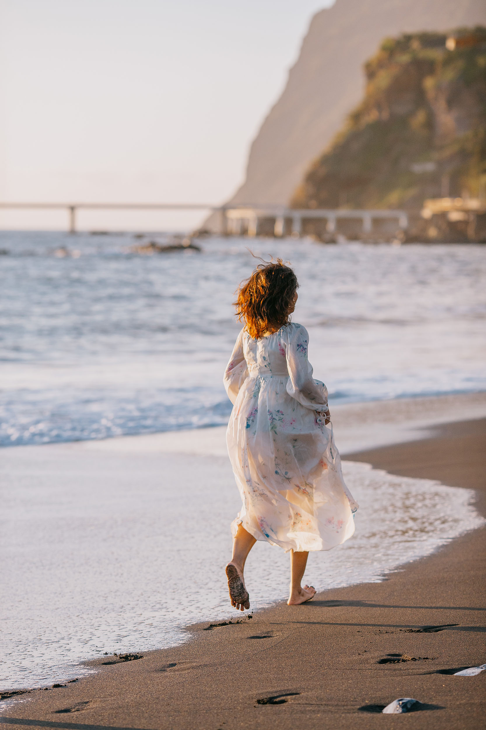Sneha running along Praia Formosa beach, white floral dress catching the wind, Funchal cliffs in the distance, Madeira