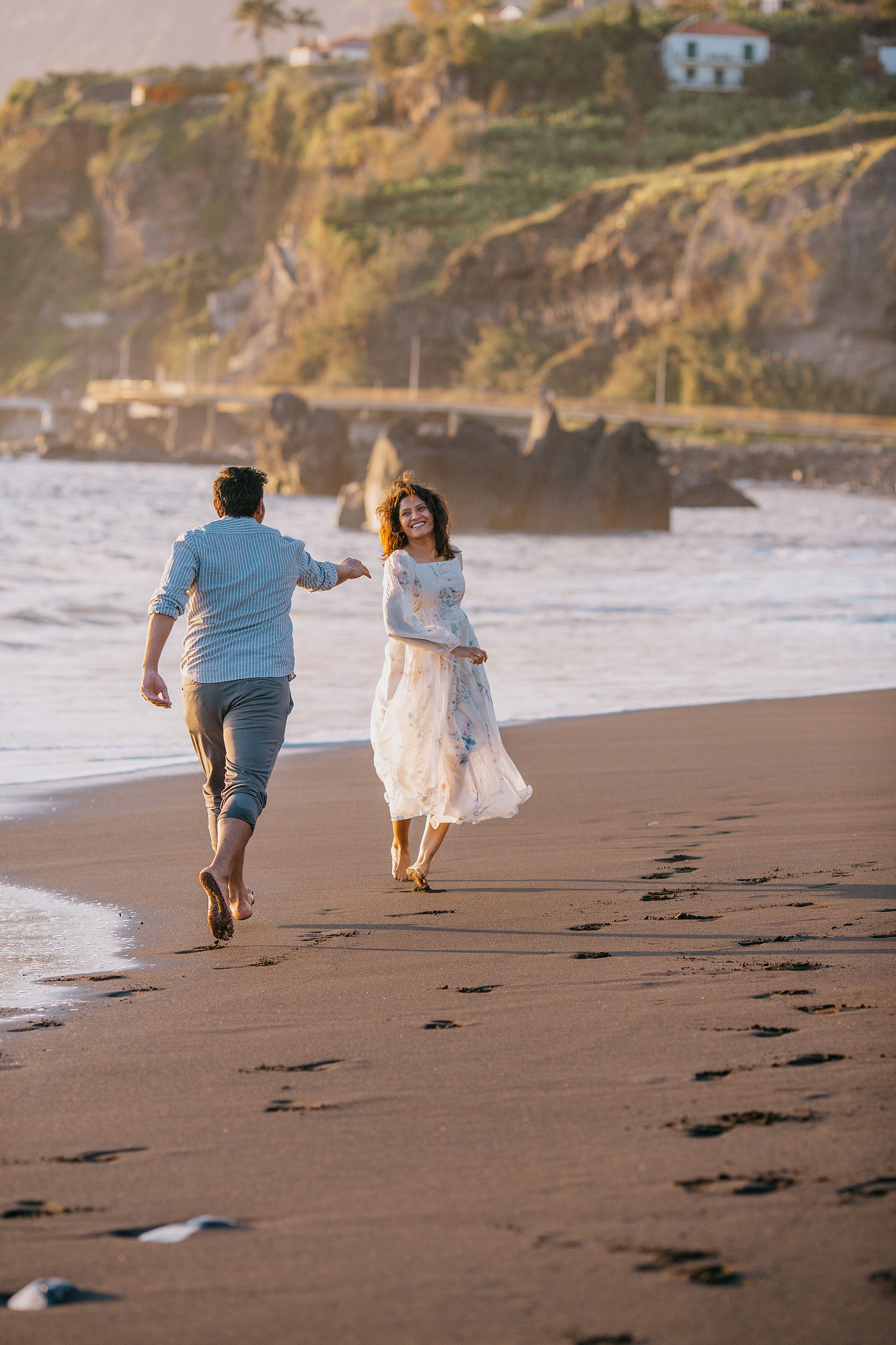 Sneha running along black sand Praia Formosa beach pulling Saket behind her, looking back laughing, Funchal cliffs behind, Madeira