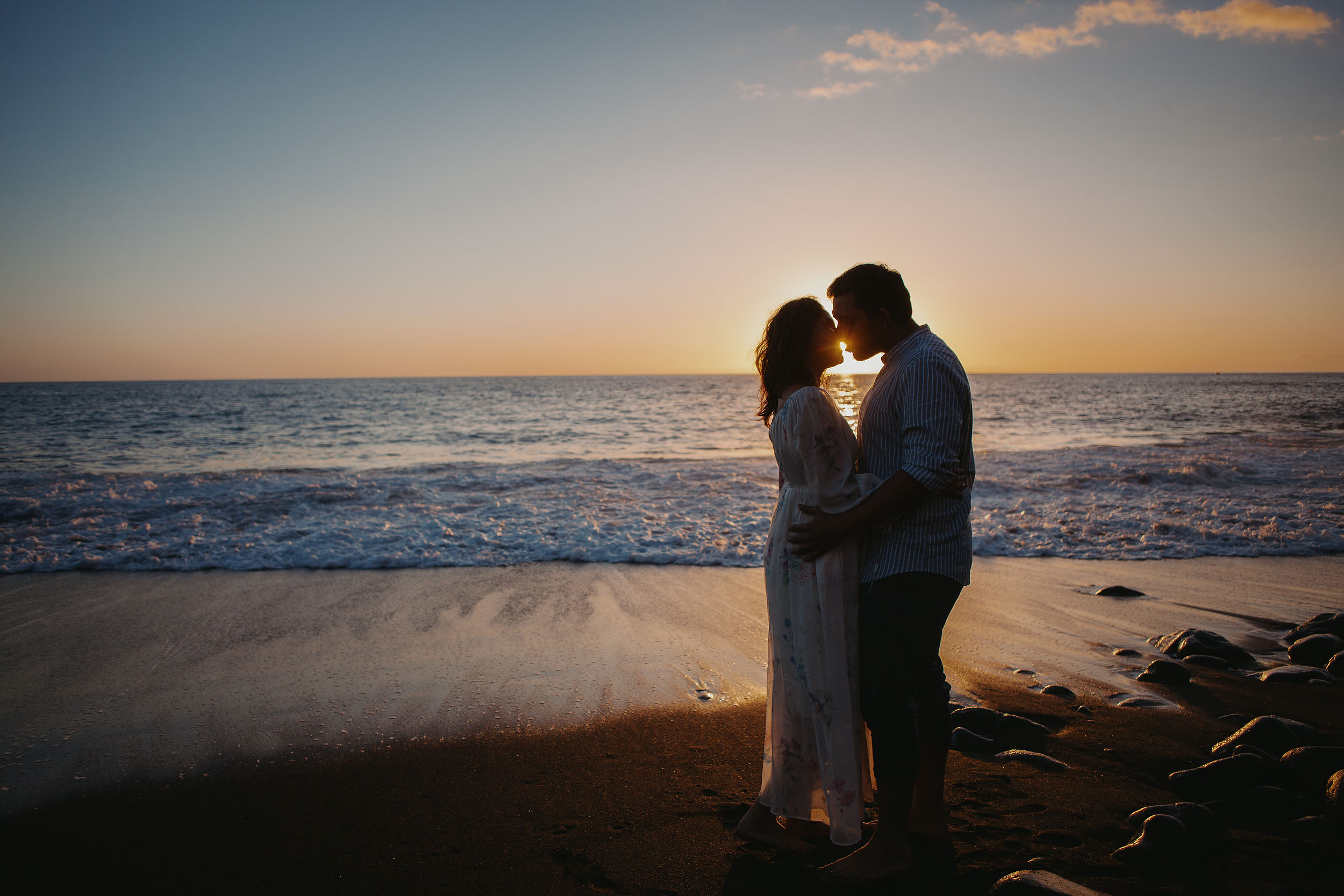 Couple silhouette kissing at sunset at Praia Formosa, Atlantic horizon glowing orange behind them, volcanic rocks at their feet, Madeira