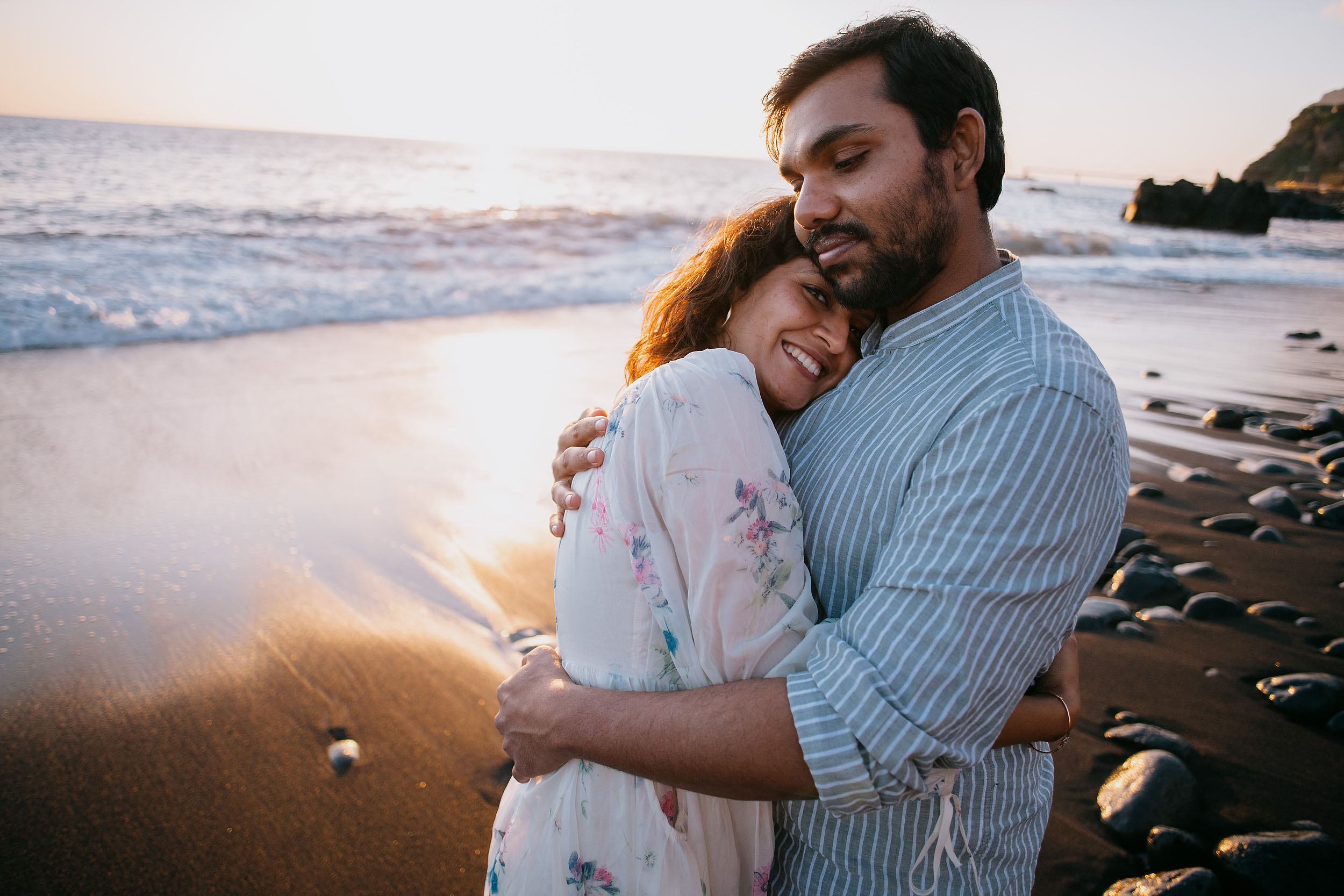 Sneha resting her head on Saket's chest eyes closed smiling, both embracing on the black sand shore at Praia Formosa, Madeira