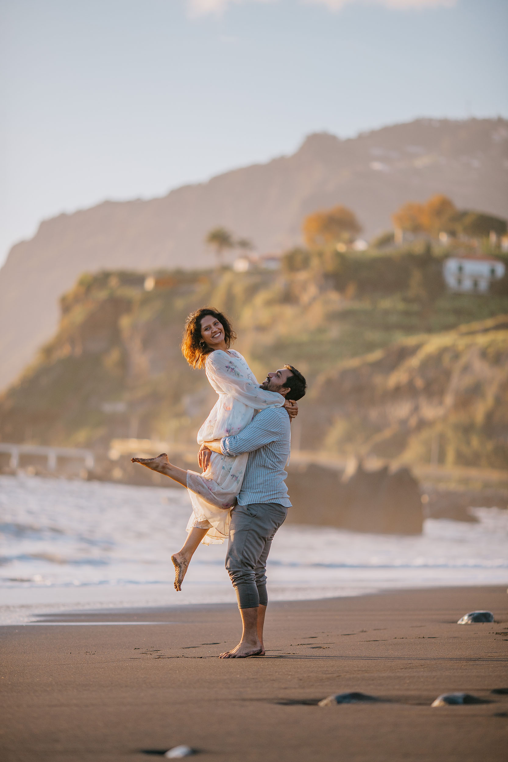 Saket lifting Sneha off the ground as she laughs with head thrown back, cream dress and denim shirt, green Funchal headland with white houses behind, Praia Formosa, Madeira
