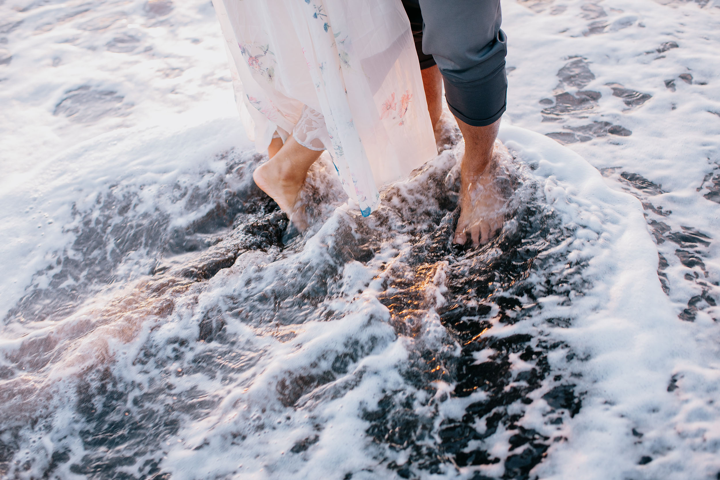 Barefoot couple standing on wave-washed volcanic rocks at Praia Formosa, Atlantic surf swirling around their feet, Madeira