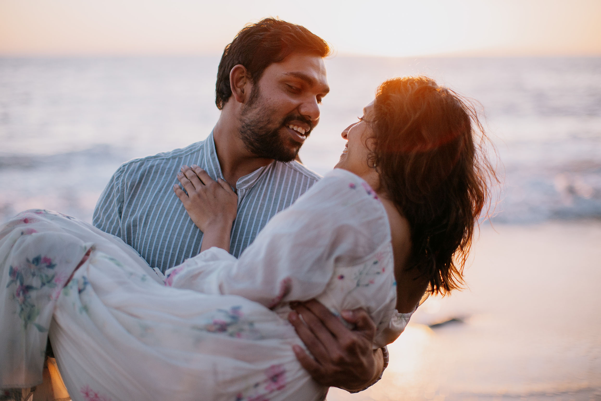 Saket laughing while carrying Sneha on Praia Formosa beach, golden sunset light over the Atlantic, Funchal, Madeira