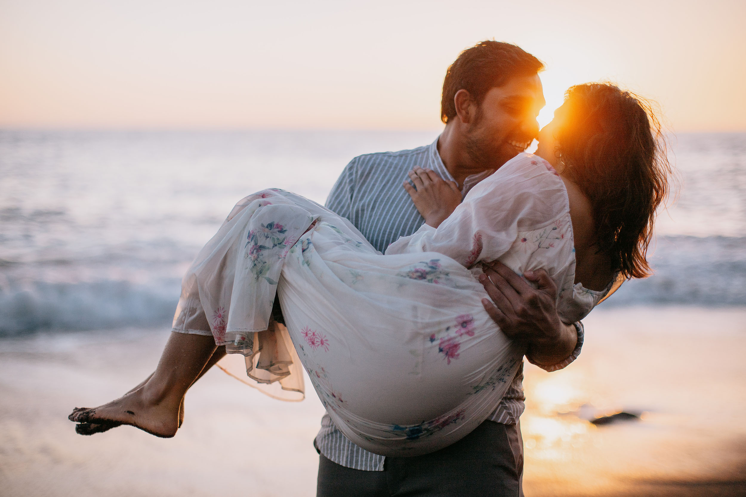 Saket carrying Sneha at sunset on Praia Formosa beach, sun flare between their faces, Atlantic behind, Funchal, Madeira