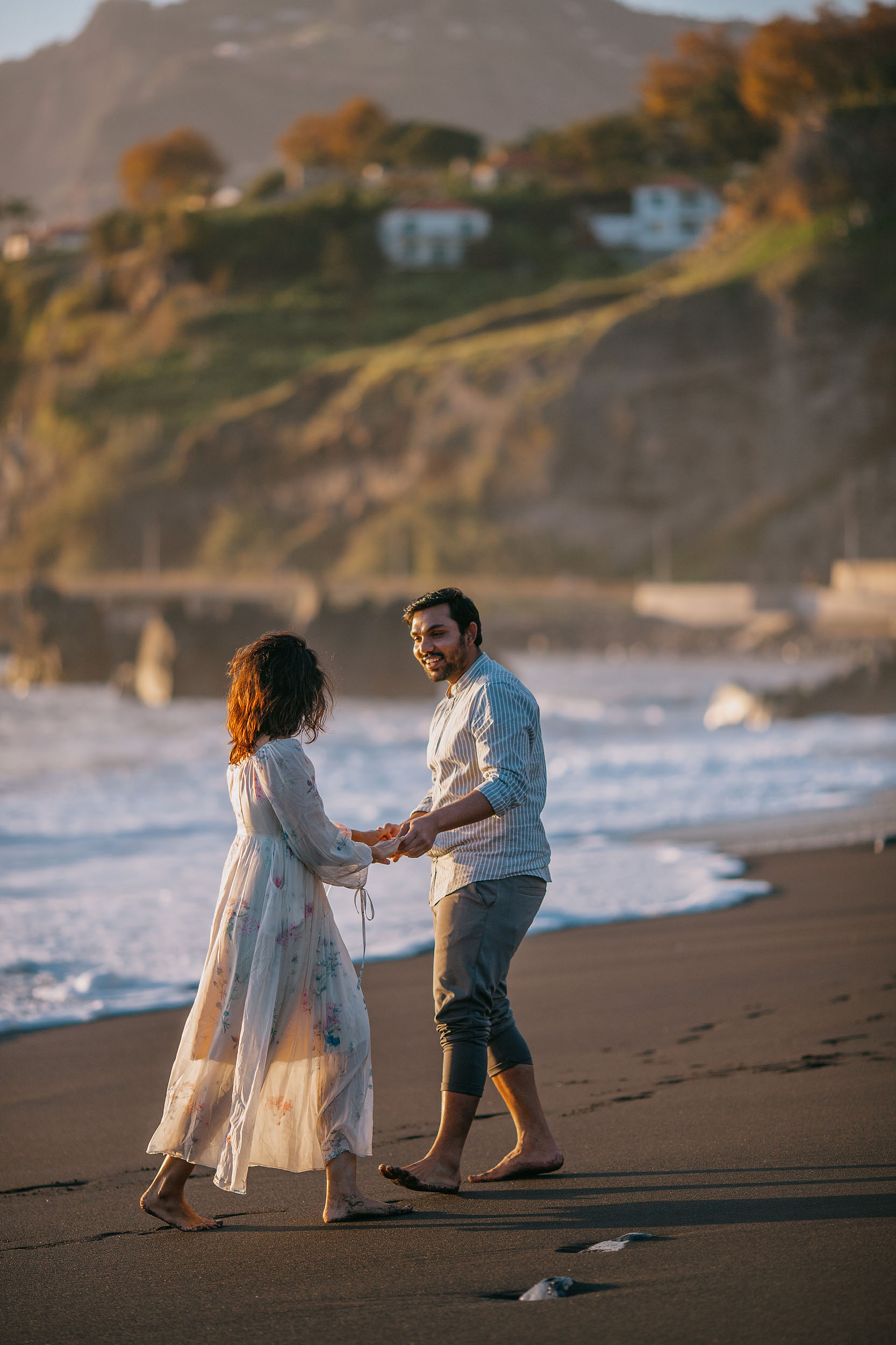 Couple holding both hands on Praia Formosa's black sand at golden hour, floral cream dress and striped shirt, green cliff and white houses behind, Madeira