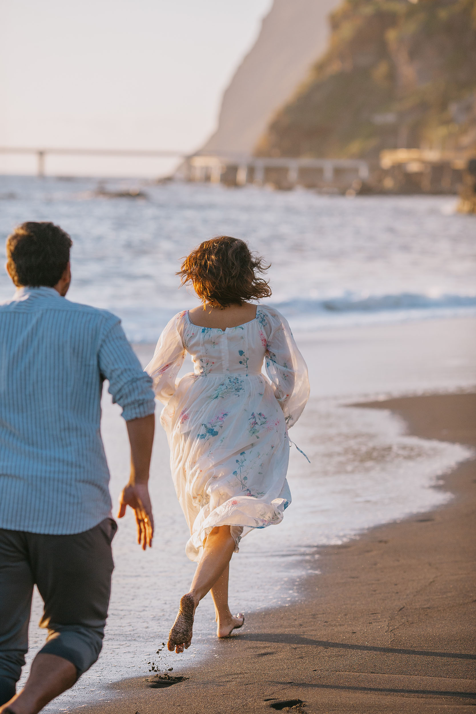 Sneha running barefoot along Praia Formosa shoreline ahead of Saket, floral dress flying, Funchal bridge and cliffs in the distance, Madeira