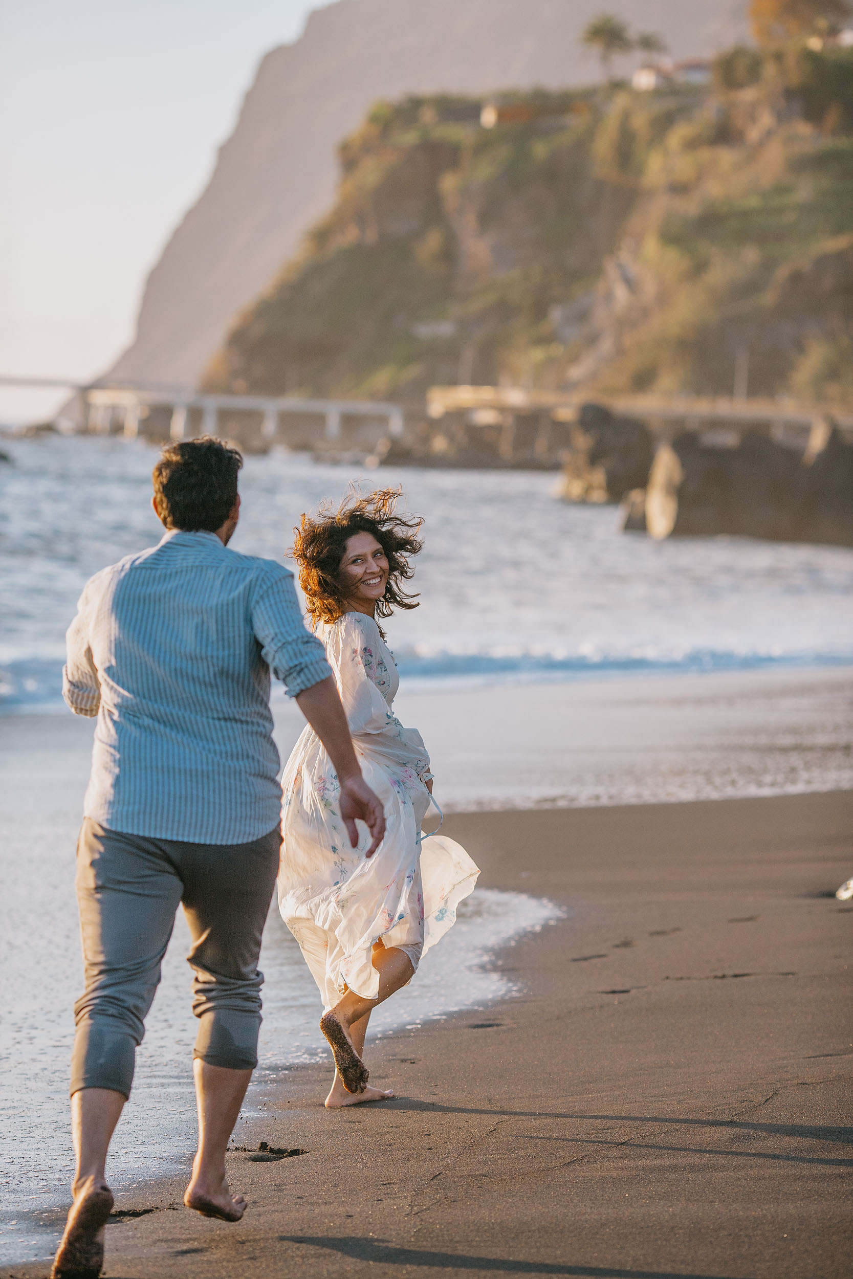 Sneha running along Praia Formosa beach laughing over her shoulder, Saket following behind, Funchal cliffs and ocean, Madeira