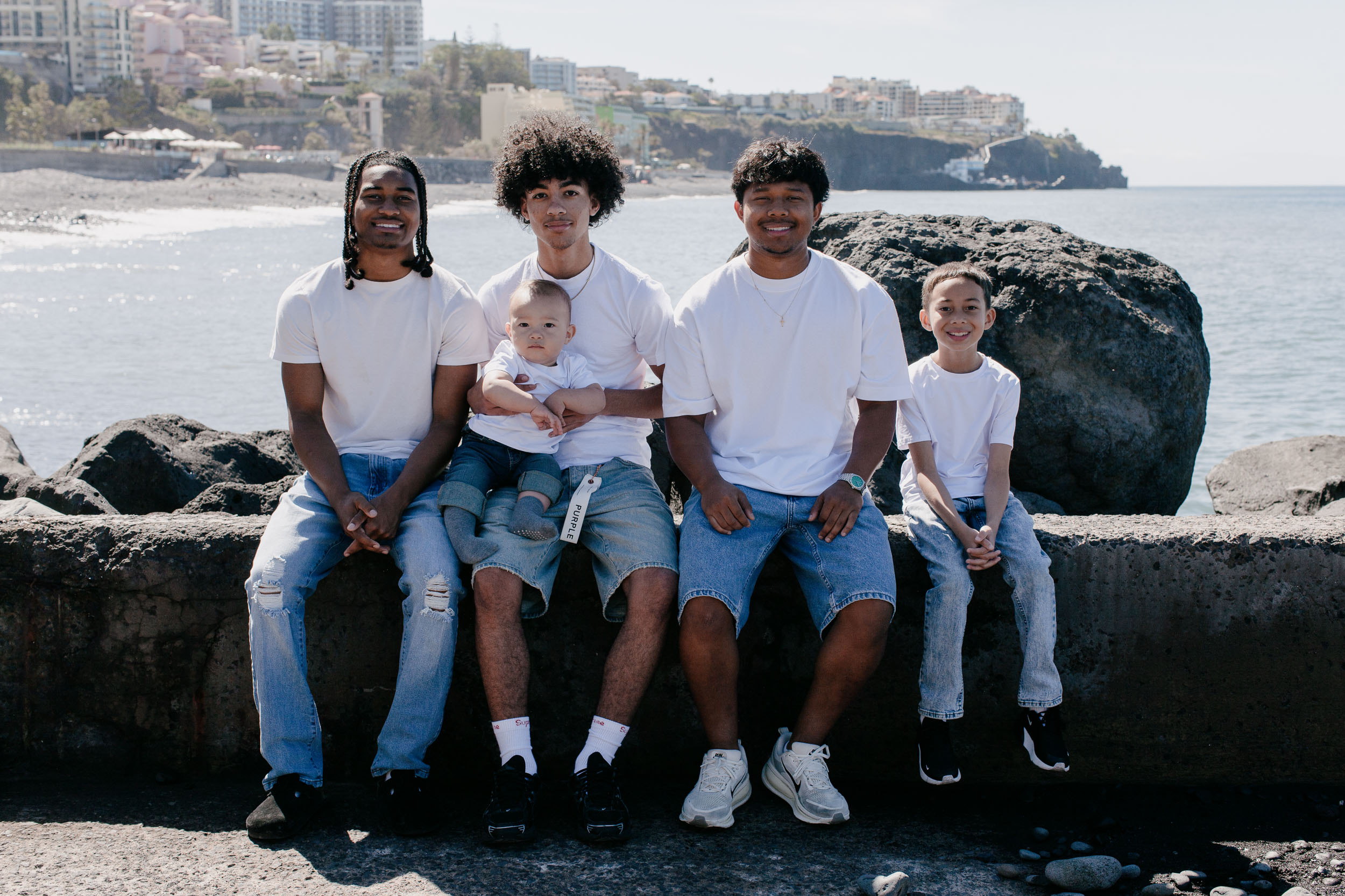 Four sons in white tees and jeans sitting on a sea wall, the eldest holding the baby, dark volcanic rock and the Funchal coastline behind, Praia Formosa, Madeira