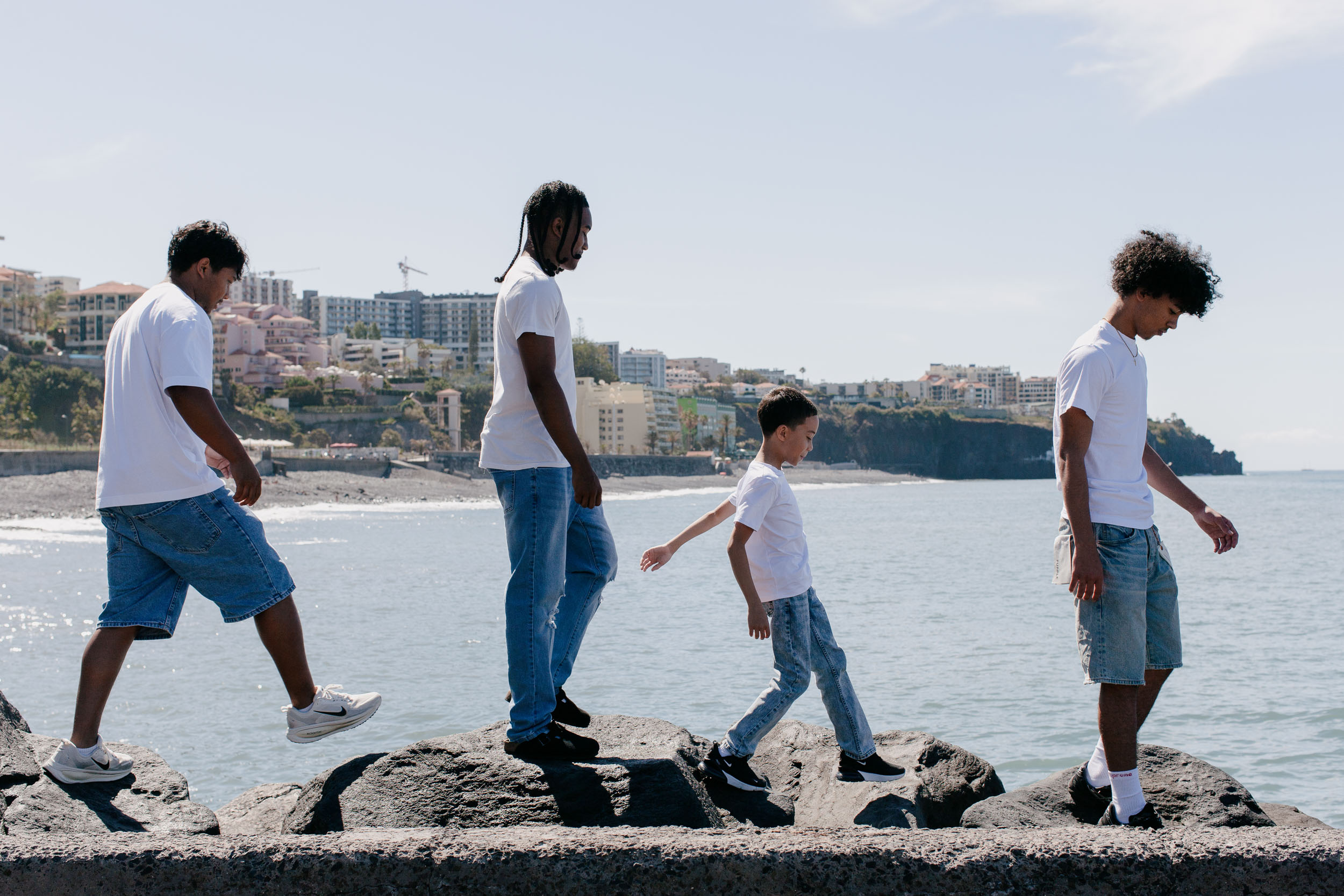 Four brothers balancing along the volcanic rocks at the water's edge, Funchal hillside and ocean behind, Praia Formosa, Madeira