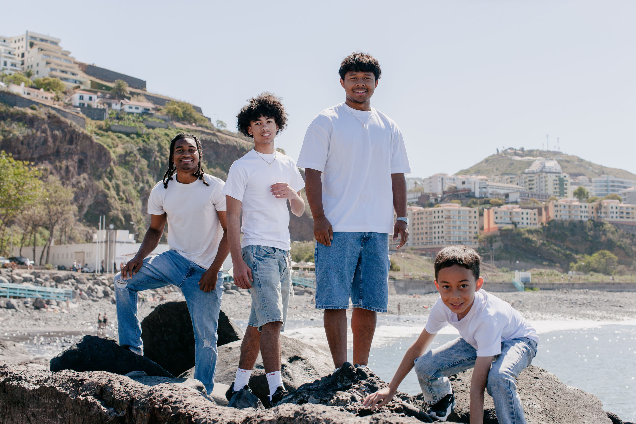 Four brothers on the rocks, three standing and the youngest crouching, Funchal hillside and ocean behind, Praia Formosa, Madeira