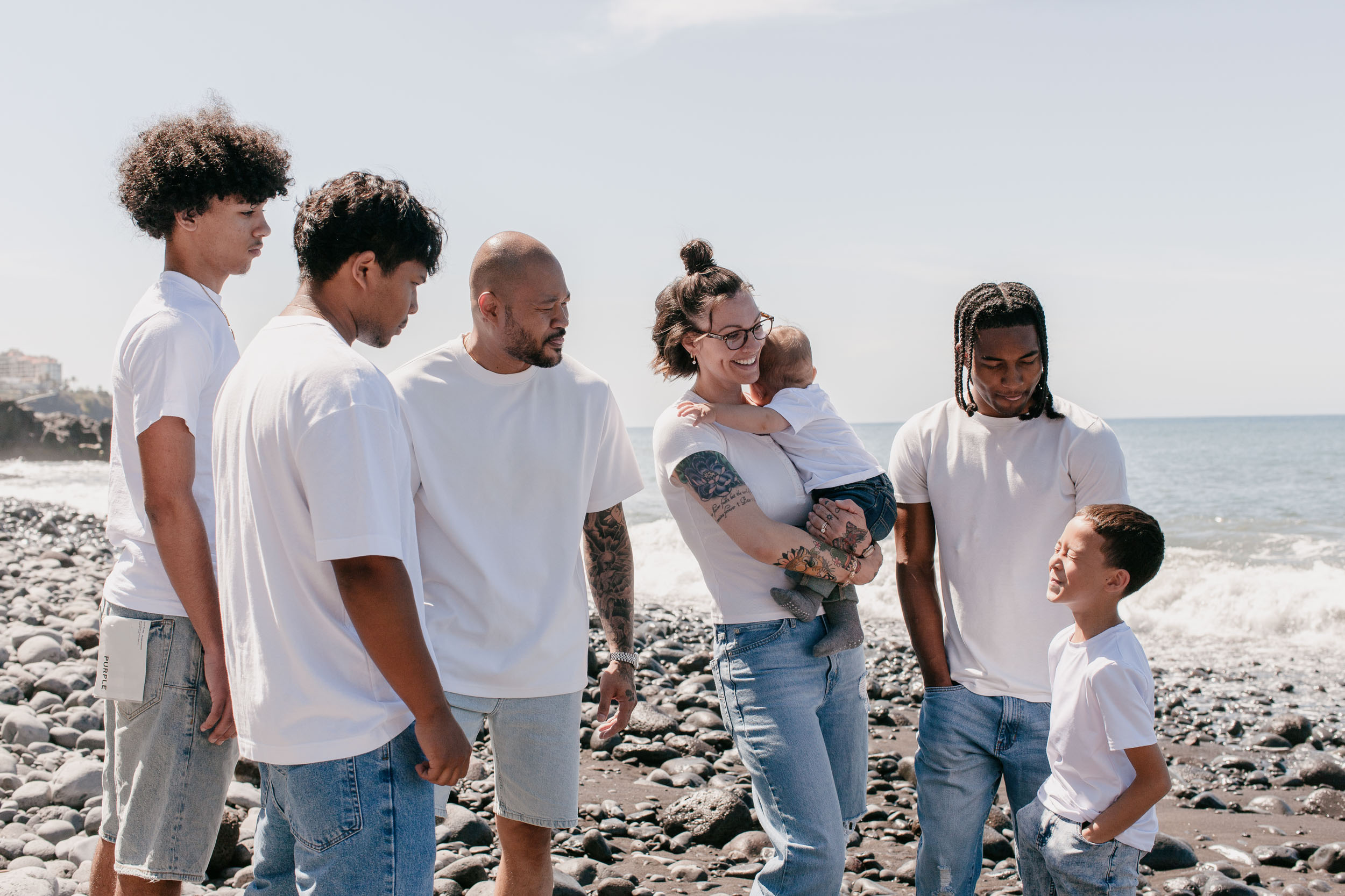 Family of seven on the pebble beach, everyone turned toward the mother and the laughing baby, the Atlantic behind, Praia Formosa, Madeira