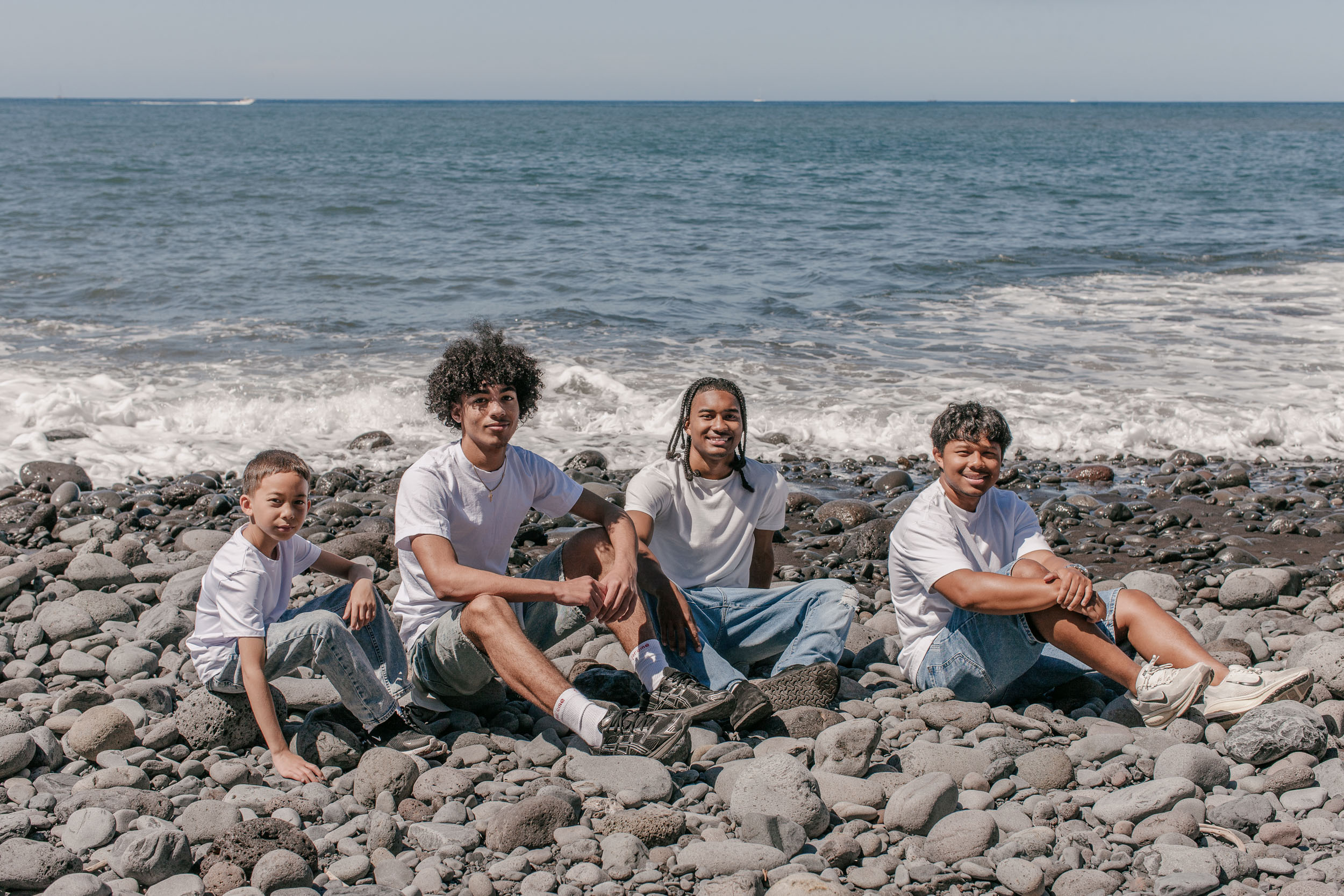 Four brothers sitting together on the pebbles at the water's edge, ocean and horizon behind, Praia Formosa, Madeira