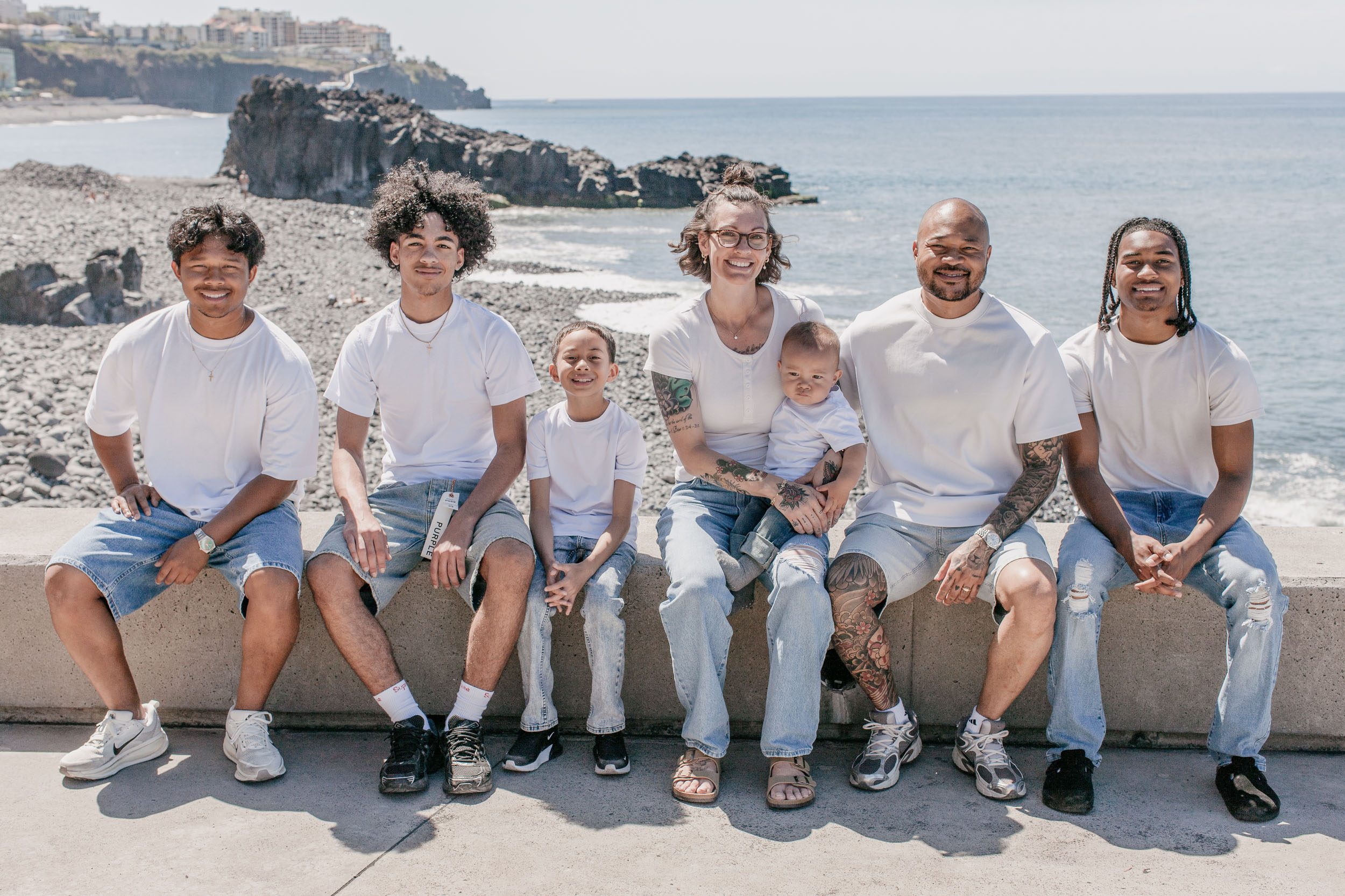 Family of seven sitting in a row on the promenade wall, volcanic headland and the Atlantic behind, Praia Formosa, Madeira