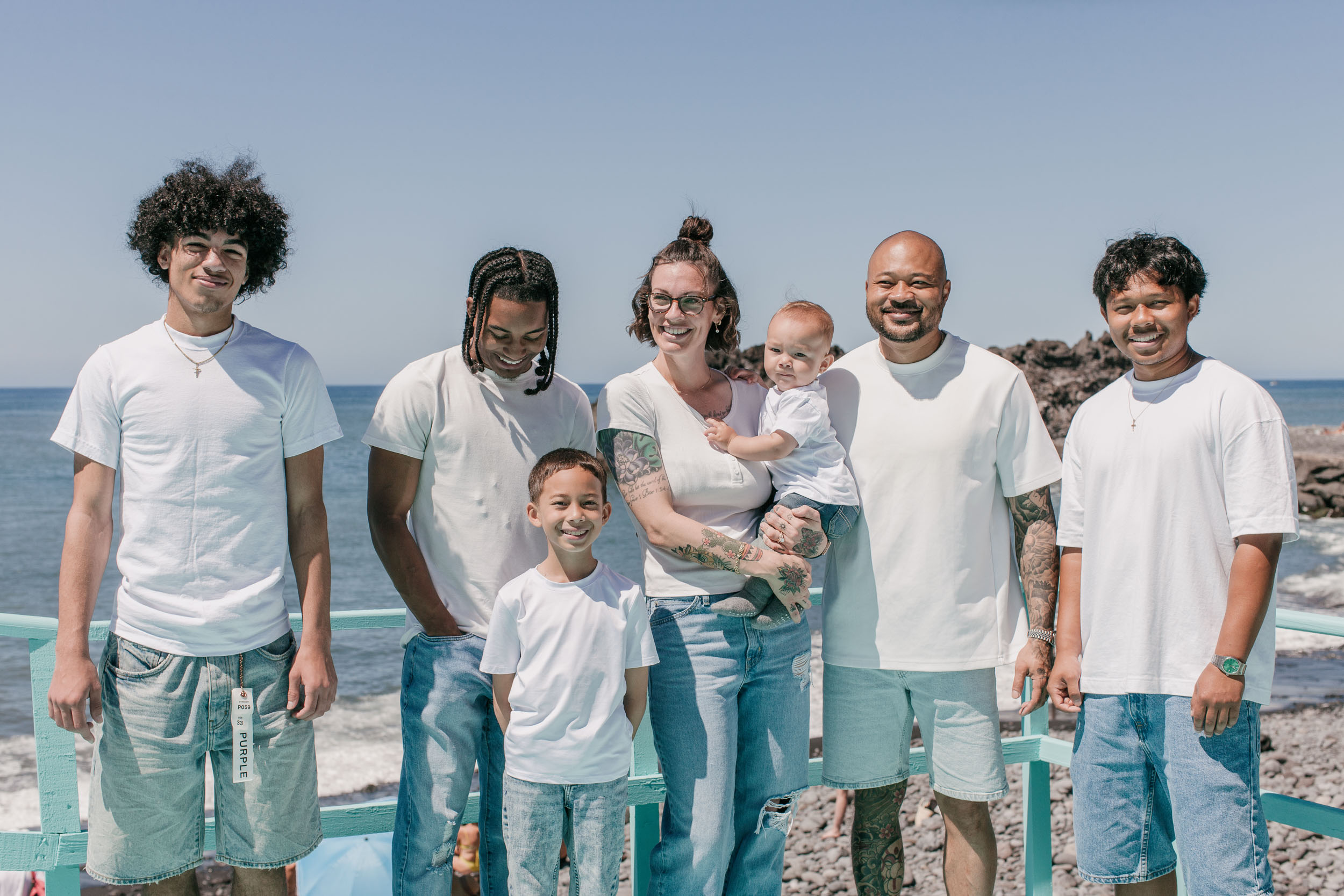 Family of seven behind the turquoise promenade railing, the rocky shoreline and ocean behind, Praia Formosa, Funchal, Madeira
