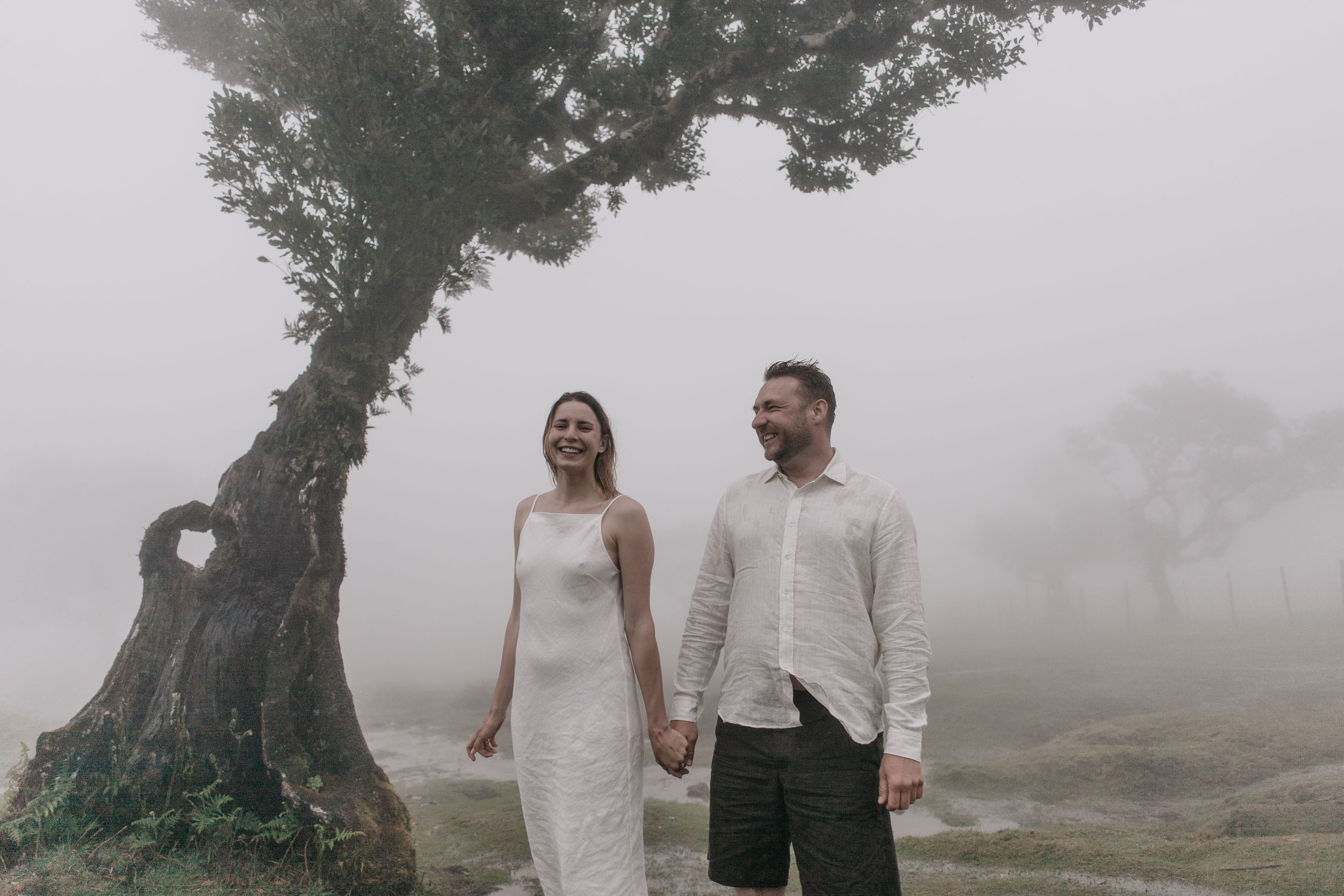 Couple in white linen under ancient laurel trees in dense afternoon fog, Fanal Forest, Madeira