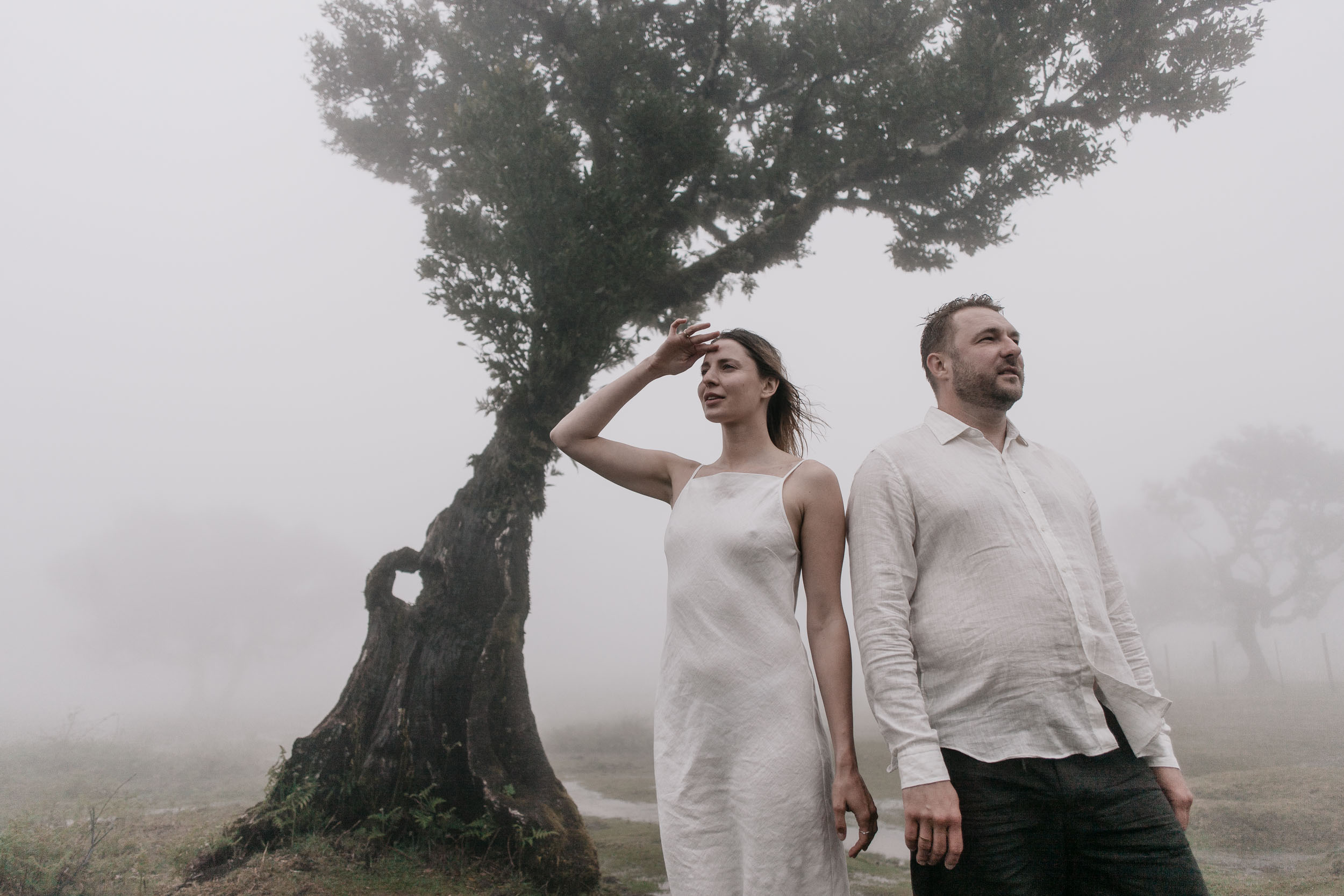 Couple stand back to back under a curved ancient laurel, her hand raised to her brow, white linen against grey mist, Fanal Forest, Madeira