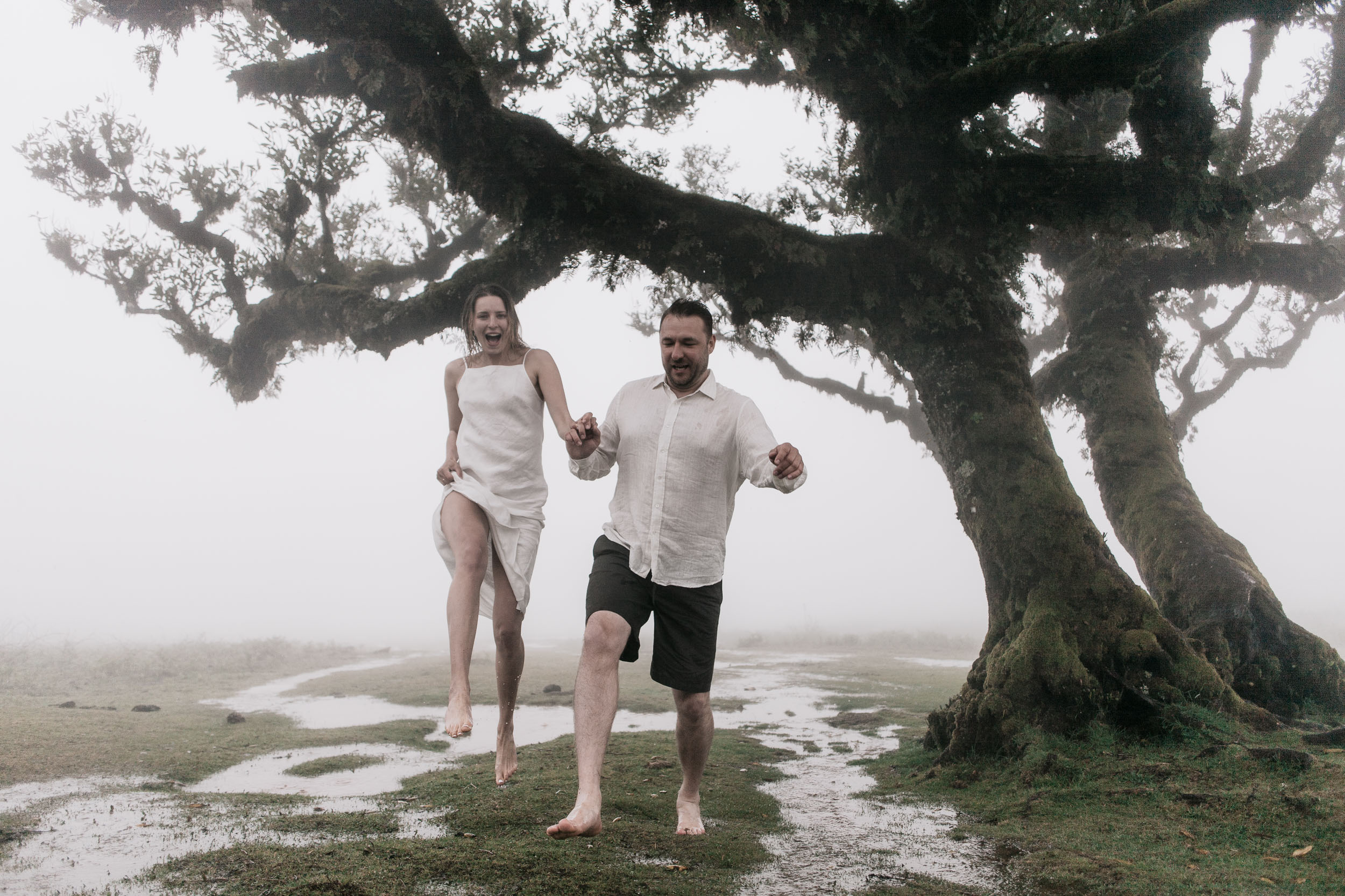Couple in front of a centuries-old laurel, branches reaching across the frame, heavy fog, Fanal Forest, Madeira