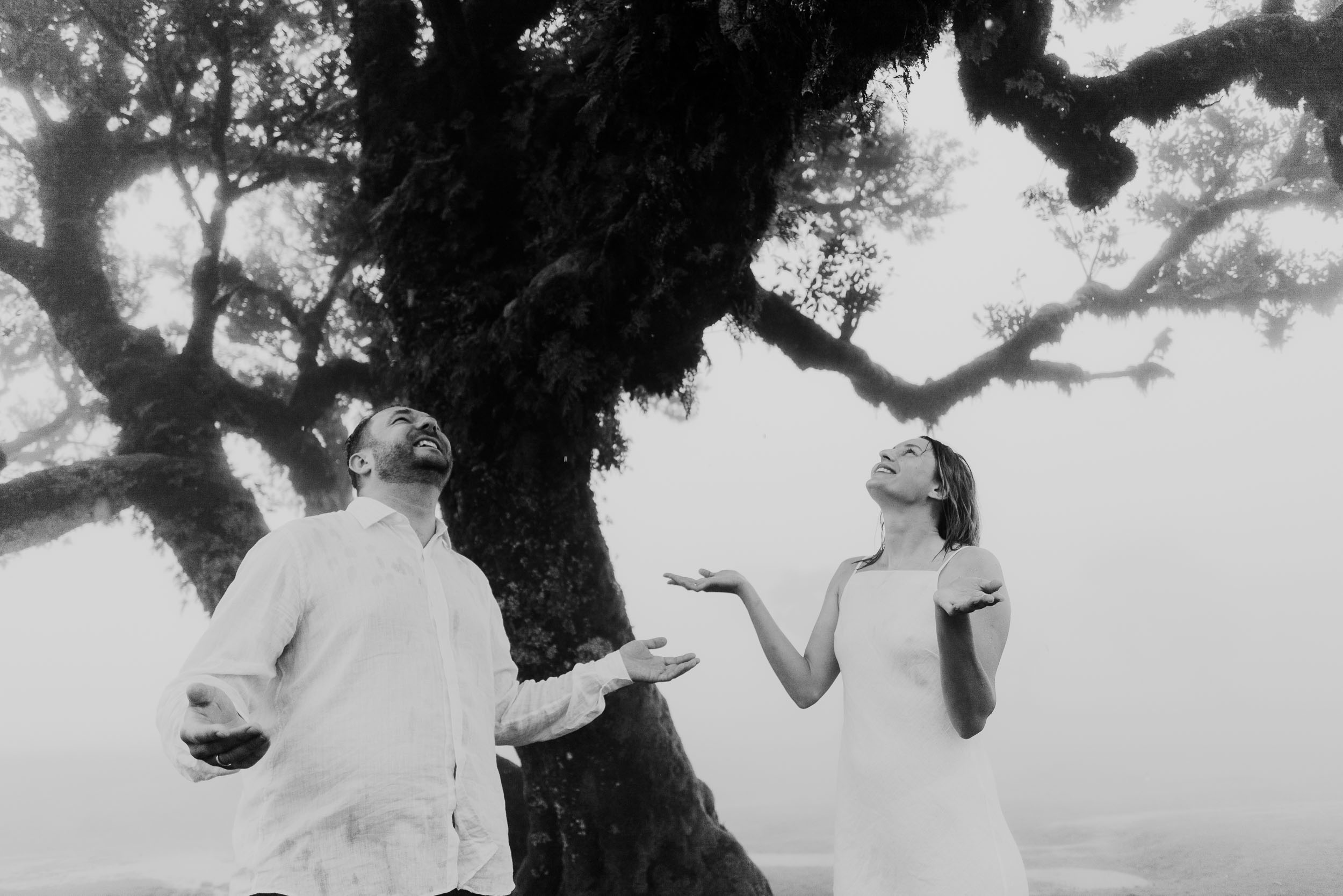 White-linen couple by a moss-covered trunk in the fog of Fanal Forest, Madeira