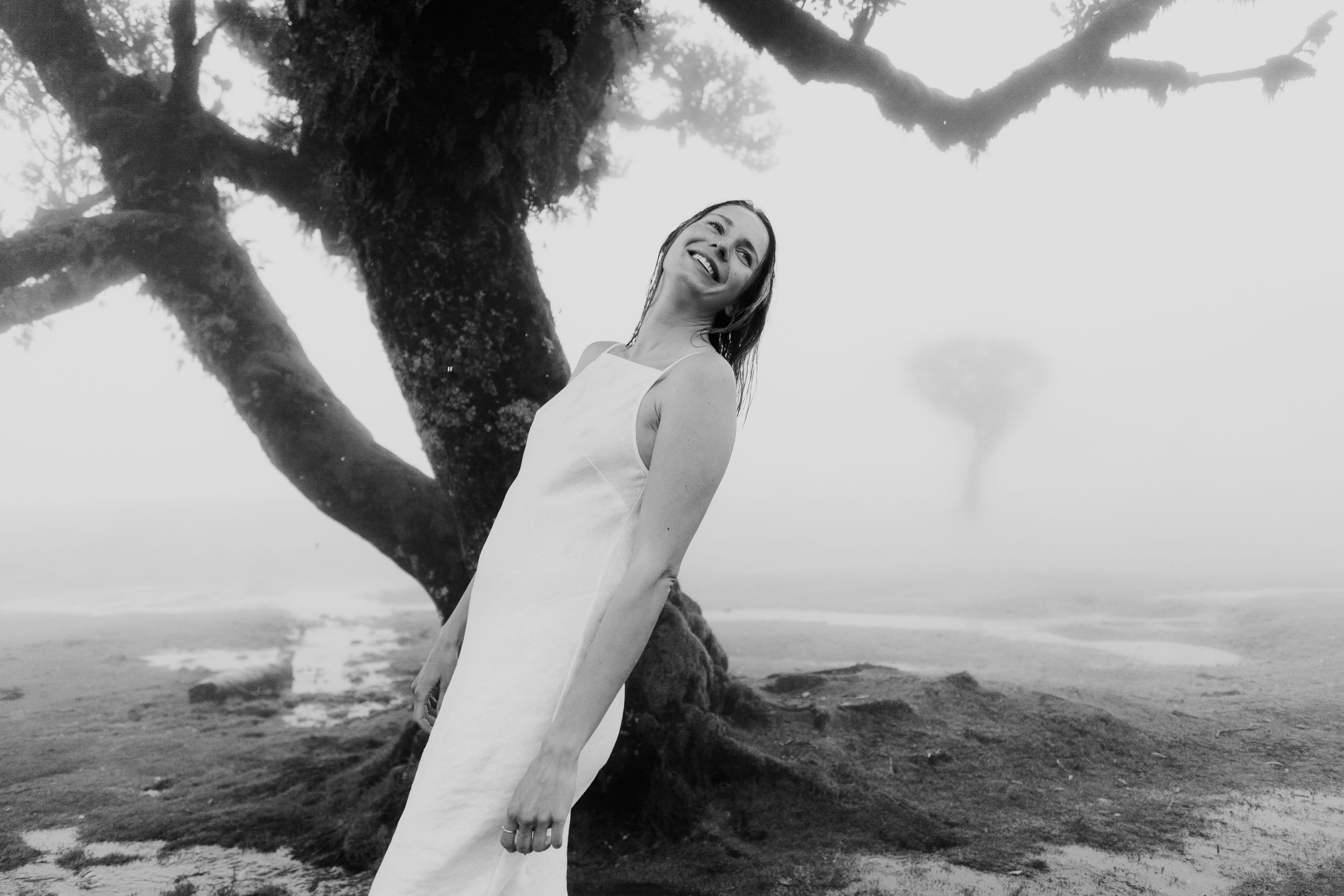 Woman in a white linen dress stands solo under twisted laurel branches, arms raised, dense fog behind, Fanal Forest, Madeira
