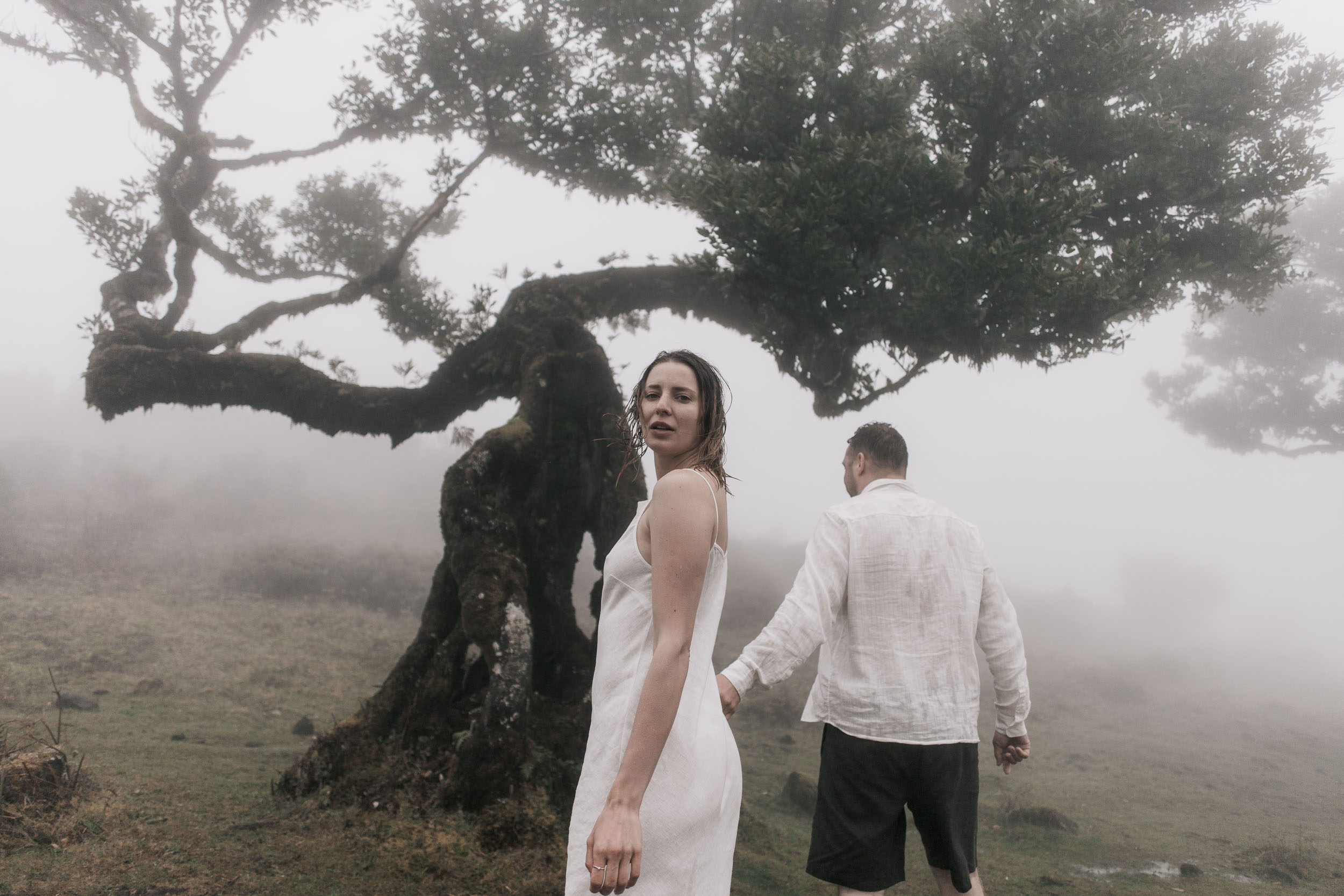 Man kneels and presses his face to her stomach by a moss-covered laurel trunk, she leans back smiling, fog behind, Fanal Forest, Madeira