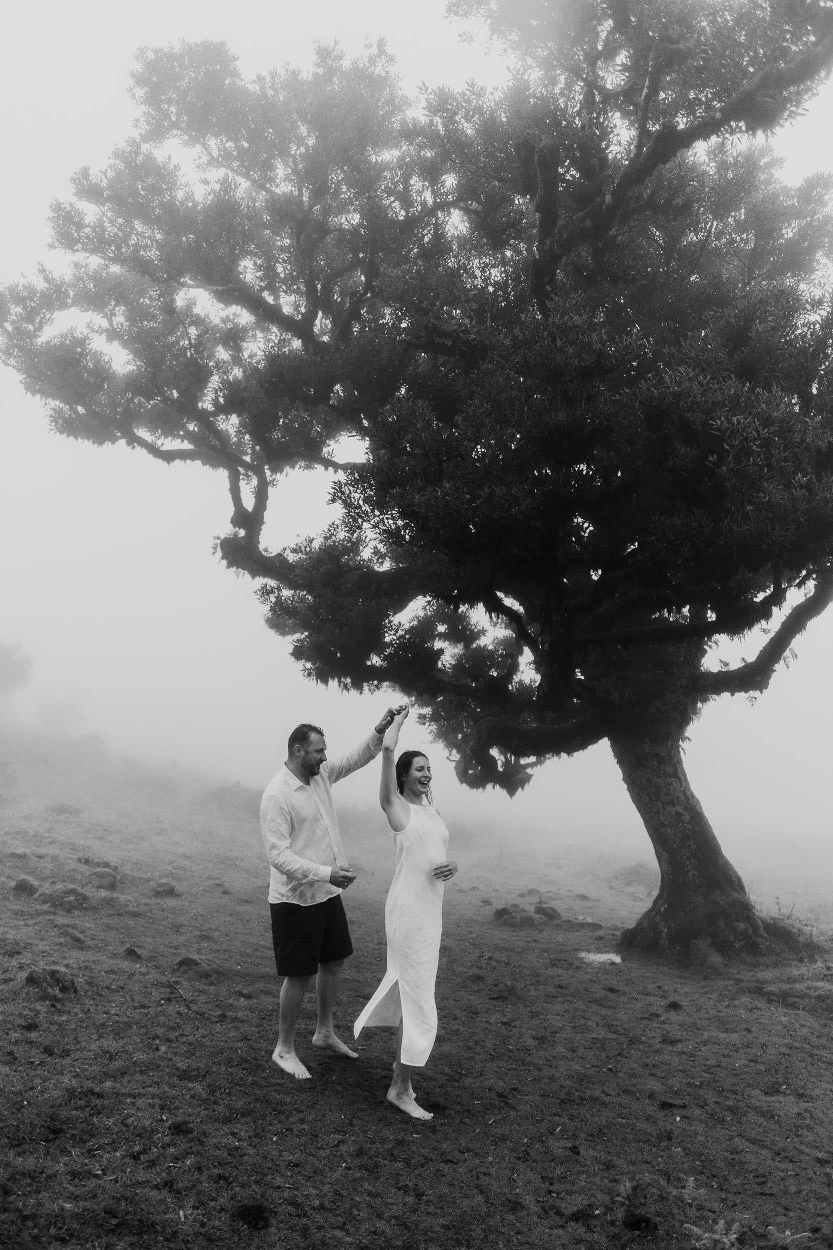 Couple dance pose under an ancient laurel tree, his arm raised guiding hers, black and white, heavy fog, Fanal Forest, Madeira