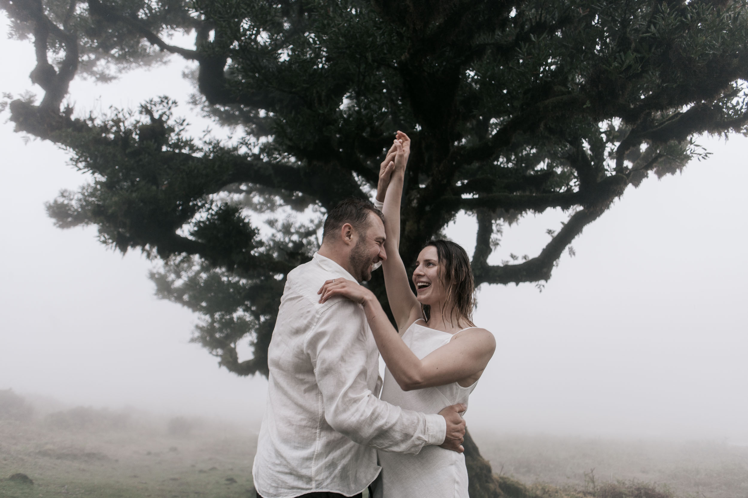 Couple in front of a centuries-old laurel, branches reaching across the frame, heavy fog, Fanal Forest, Madeira