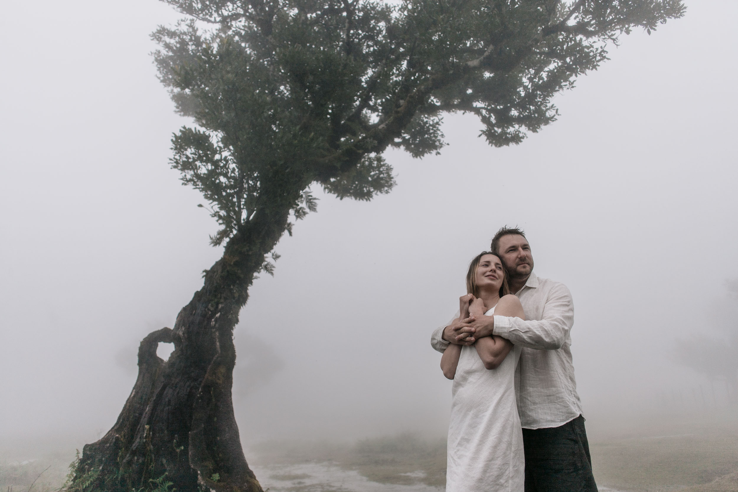 Couple stand together in front of a moss-covered ancient laurel, he holds her from behind, both in white linen, dense fog filling the background, Fanal Forest, Madeira