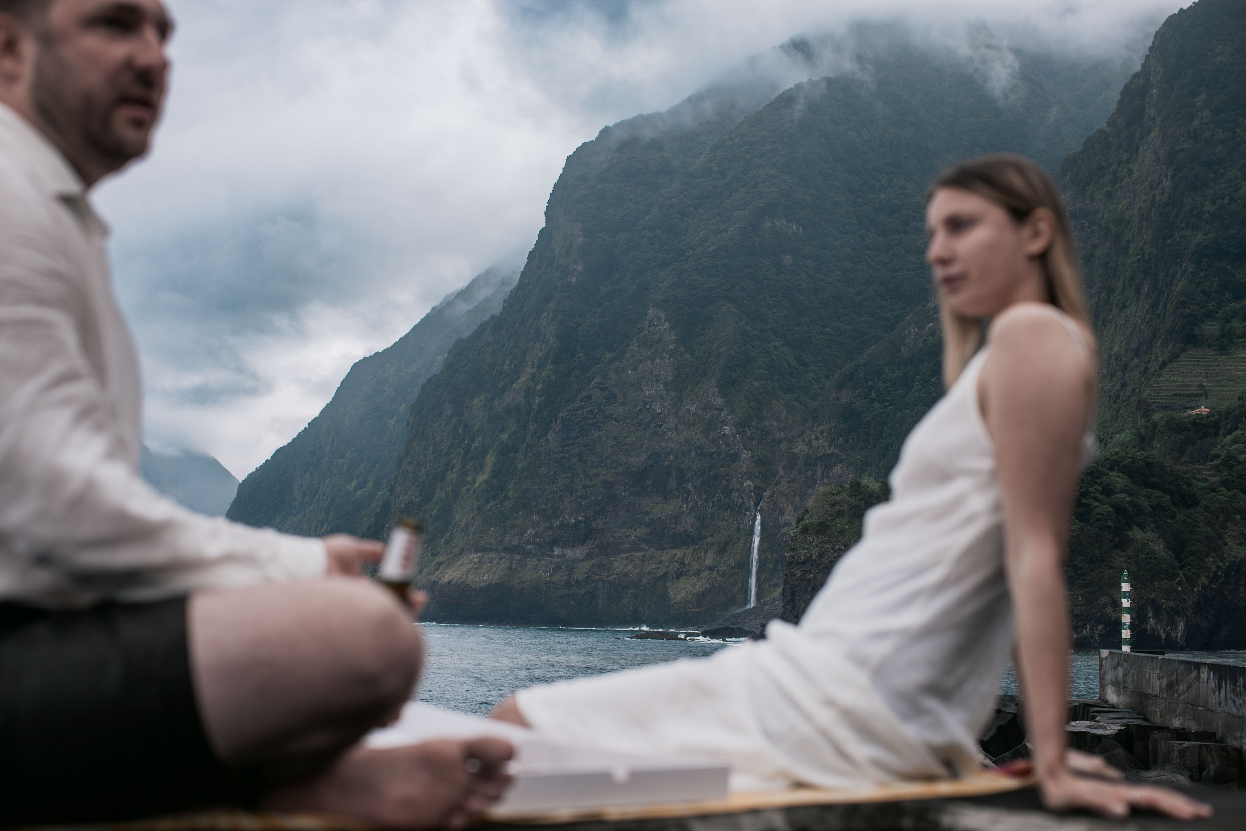 Couple in white share a pizza on a stone breakwater above the sea, lighthouse and cliffs behind, north Madeira
