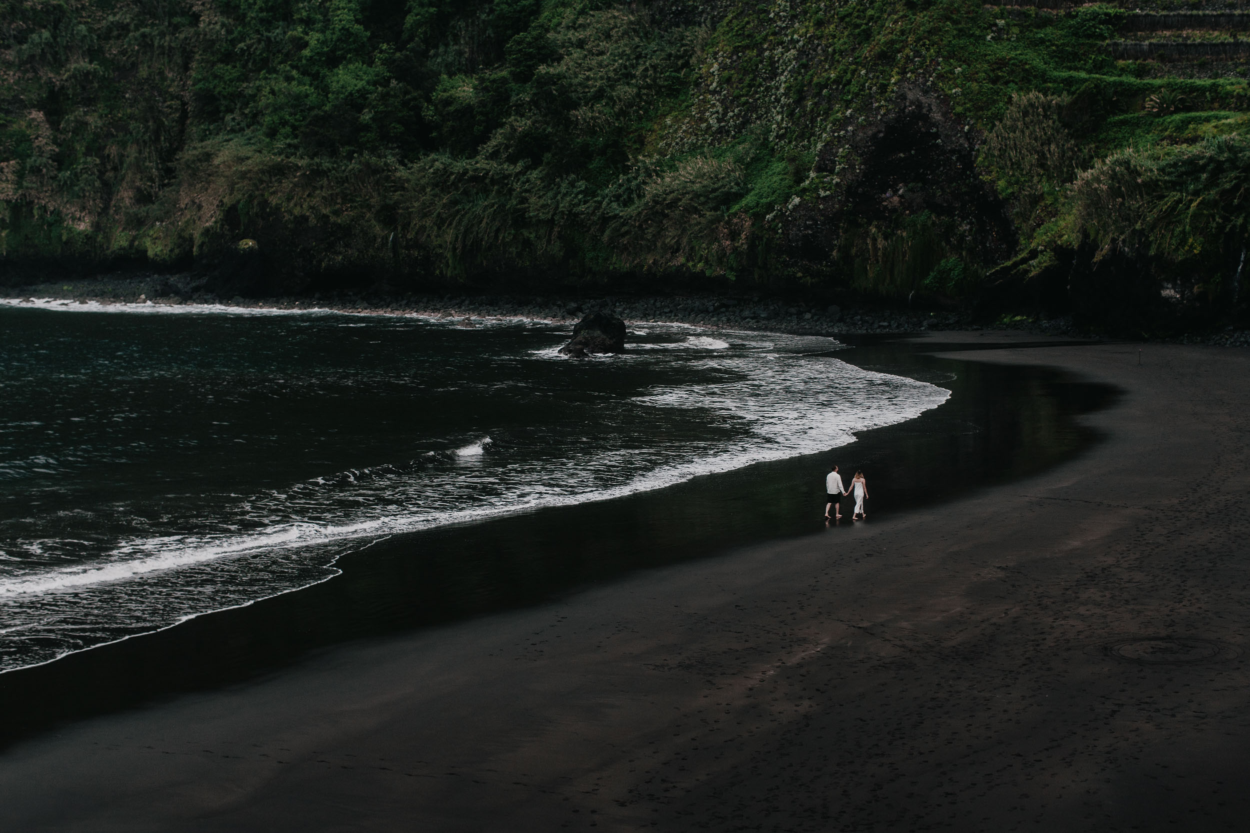 Wide landscape: tiny couple holding hands at the edge of a black sand cove, sheer green cliffs framing both sides, north Madeira