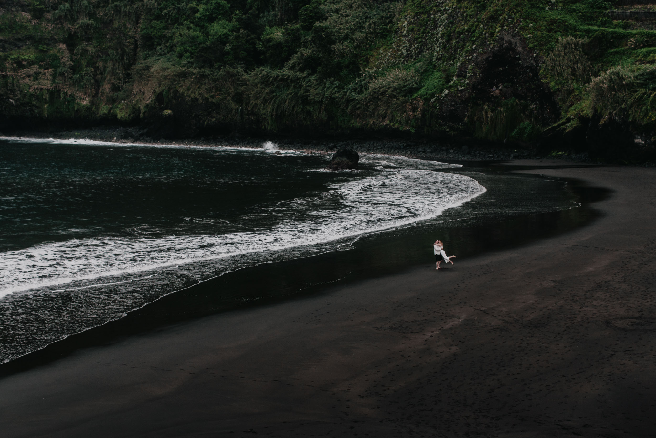 Wide pejzaze of a dark beach with the couple walking at the waterline, dramatic cliffs behind, north Madeira