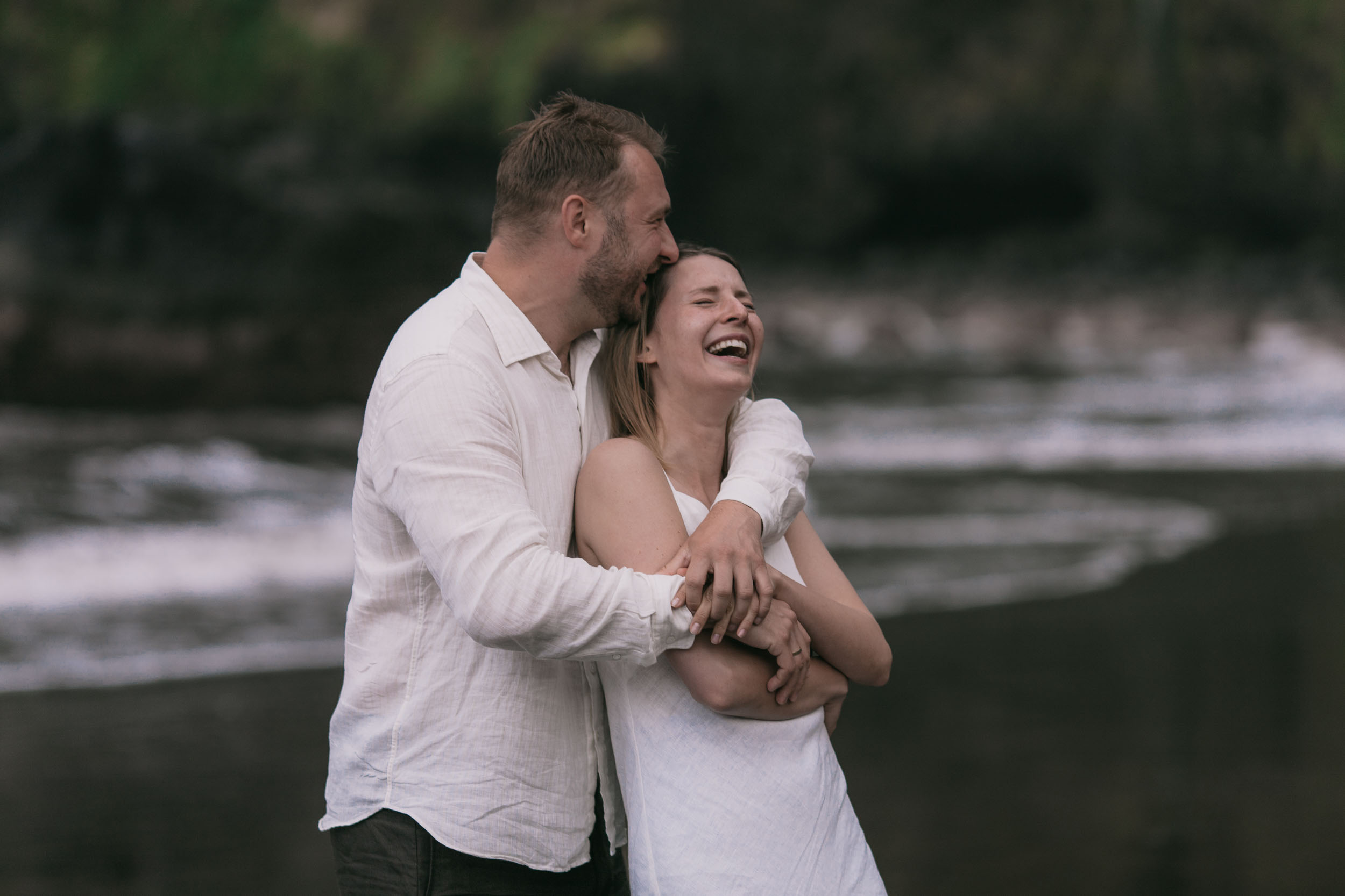 Couple wrapped together on a dark beach, laughter on her face, north Madeira coast