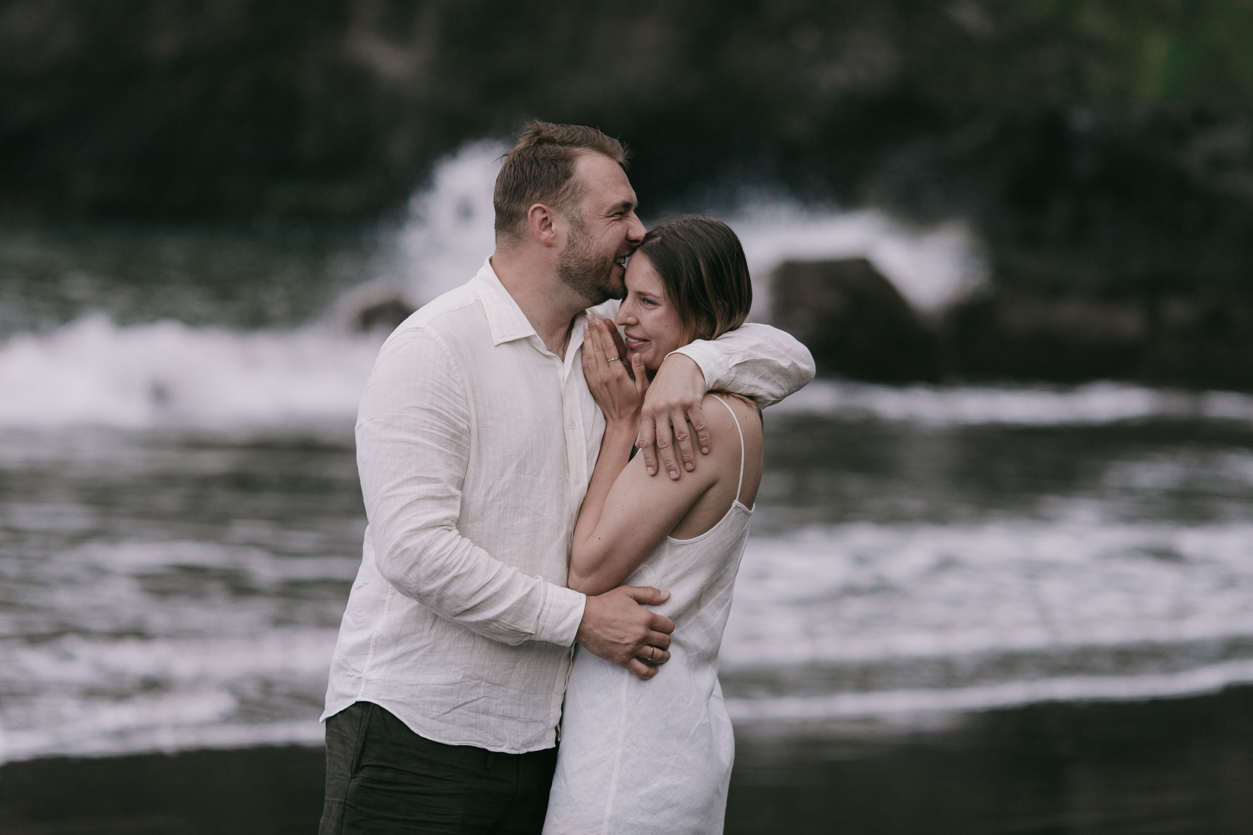 Close embrace on a black sand beach, she rests her face on his shoulder smiling, ring visible on her finger, white linen, Seixal Beach area, Madeira