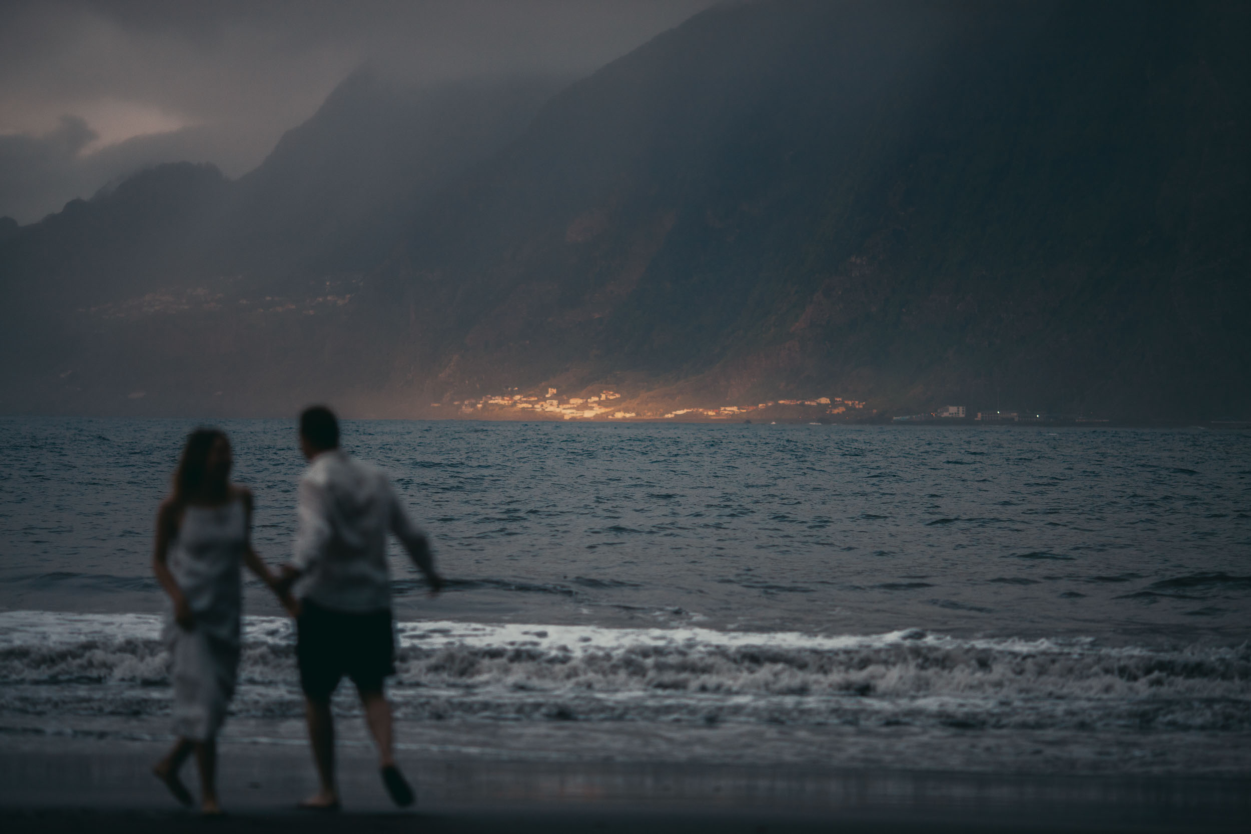 Couple walking hand in hand on a dark beach at dusk, shifted out of focus, sharp lit village across the bay on a green ridge, Madeira