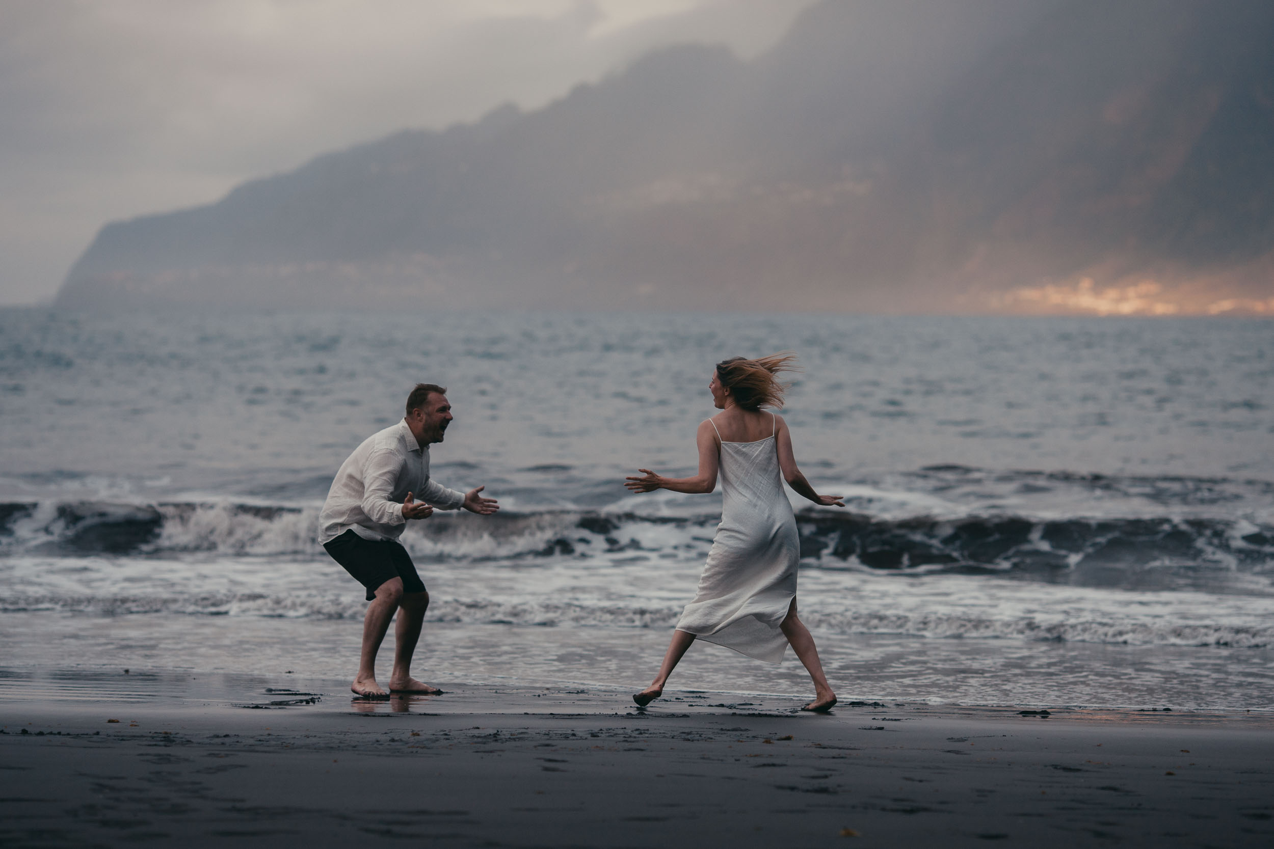 Couple running toward each other on a dark sand beach at sunset, dramatic arms outstretched, fog drifting over the cliffs, Madeira